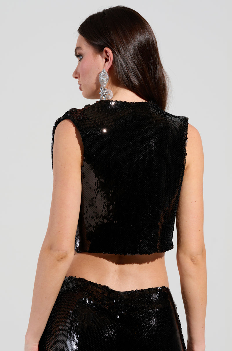 A person with long dark hair wears a sleeveless crop top from the AKIRA Label, specifically the PARTY IN THE CITY CROP TOP IN BLACK, adorned with sequin embellishments, paired with matching pants. They sport large statement earrings. The person's pose is slightly turned away, highlighting the garment's texture and shine.