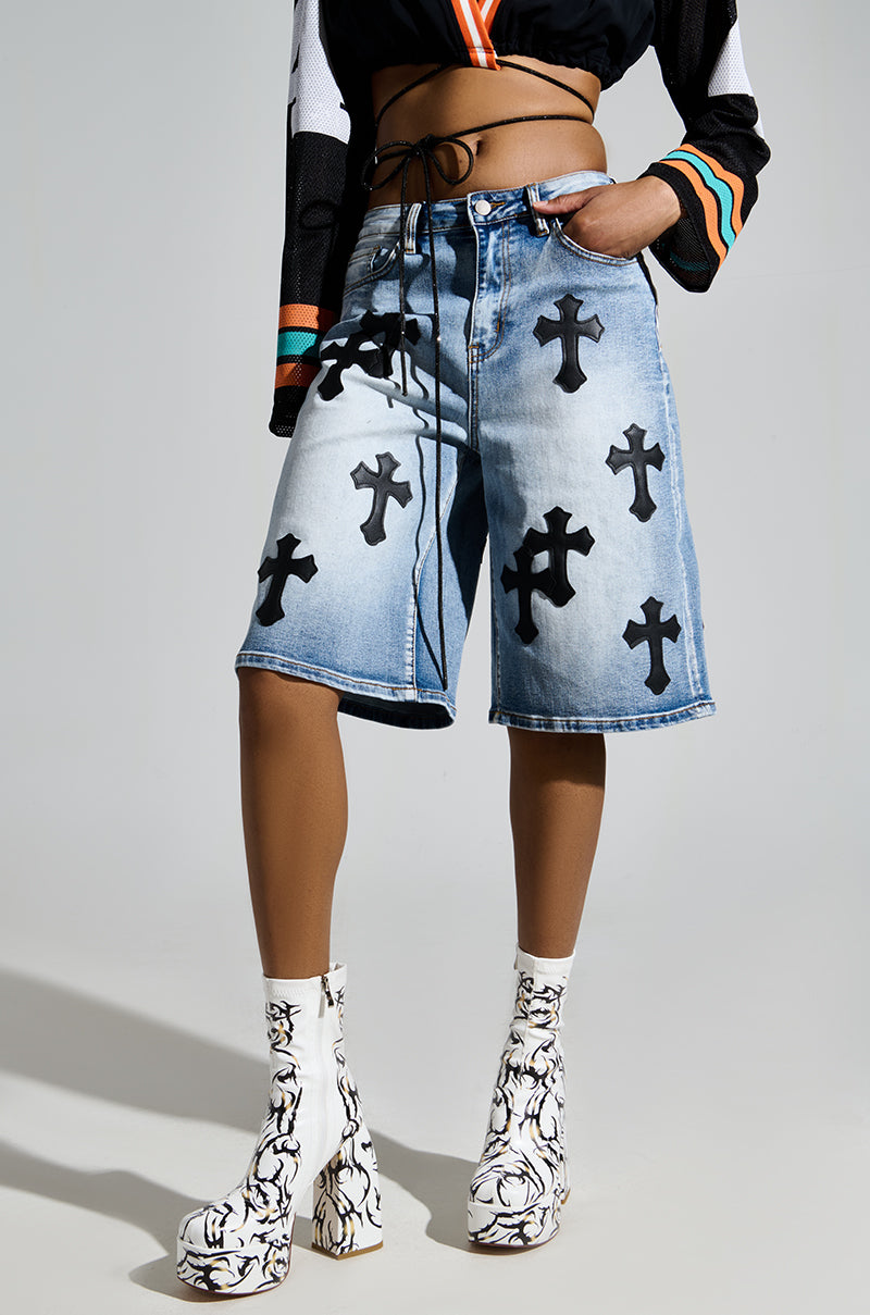 A person wears denim shorts with black cross patches, a black top with colorful sleeve accents, and the AZALEA WANG ARIADNE WHITE CHUNKY BOOTIE featuring a tattoo-inspired pattern, standing against a plain background.