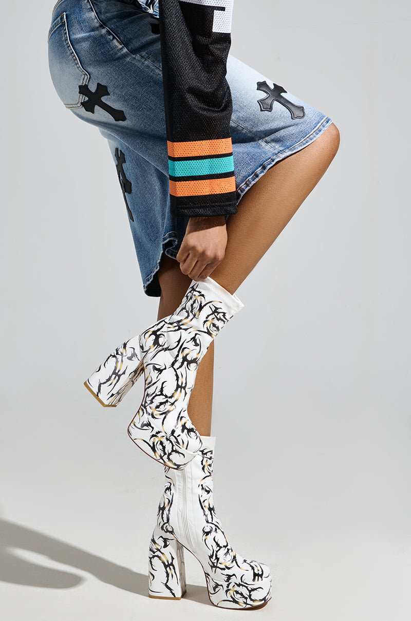A person wearing a skirt and colorful long-sleeve shirt poses with a bent leg, highlighting the AZALEA WANG ARIADNE WHITE CHUNKY BOOTIE featuring a bold black and yellow tattoo-inspired design, set against a plain grey background.
