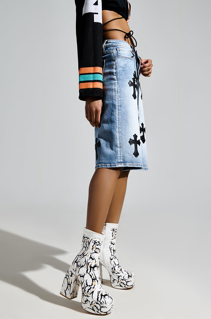 A person dons a black long-sleeve crop top and a blue denim skirt with black cross patterns, paired with AZALEA WANG ARIADNE WHITE CHUNKY BOOTIES featuring tattoo-inspired vine designs on platform soles. The background is plain gray.