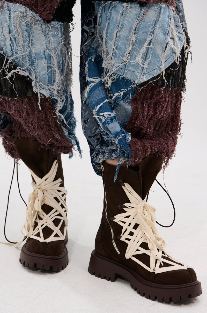A close-up of a person in blue and burgundy distressed patchwork pants, paired with AZALEA WANG ARJUN CHOCOLATE LACE BOOTIE featuring a chunky heel and oversized cream-colored laces wrapped around the boots.