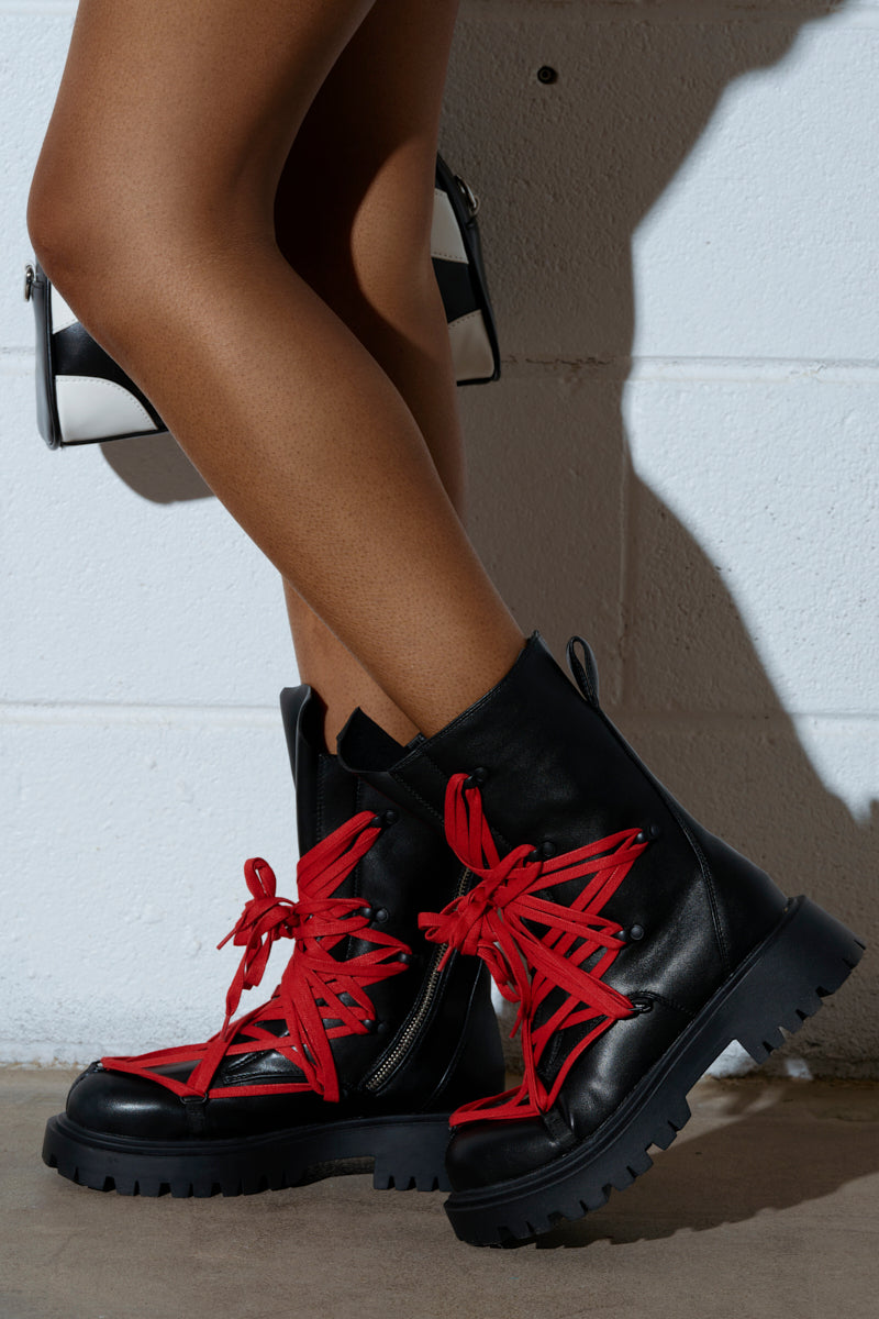 Someone wearing the AZALEA WANG ARJUN Lace Detail Combat Bootie in black with red lace stands against a white wall, casting a shadow with crossed legs. They hold a black and white bag. The vegan leather boots have side zippers and thick soles.