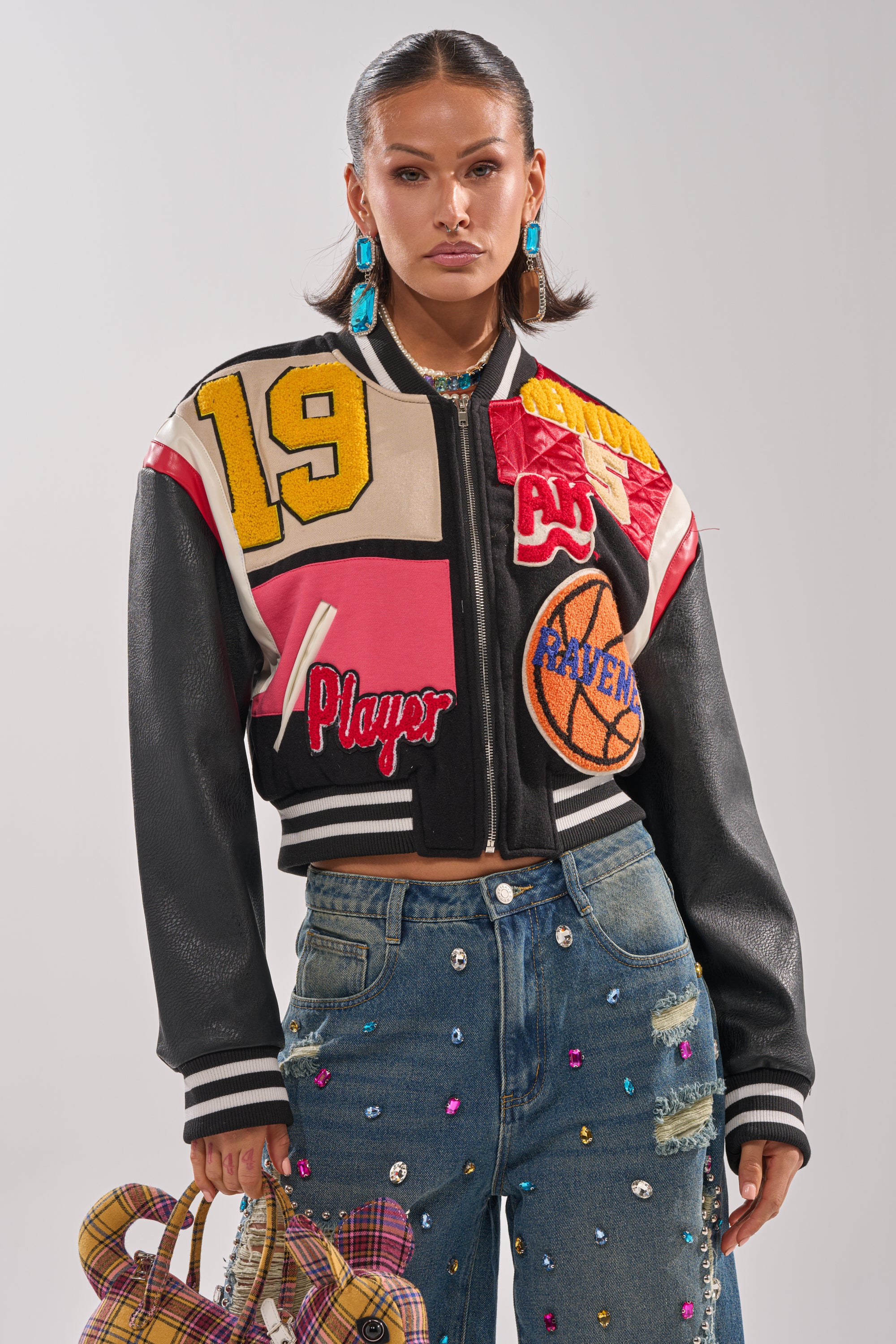 A woman wears the NEW YORK NEW YORK VARSITY BOMBER with basketball letterman patches over gem-studded high-waisted blue jeans, holds a plaid handbag, and accessorizes with bold earrings against a plain light background.
