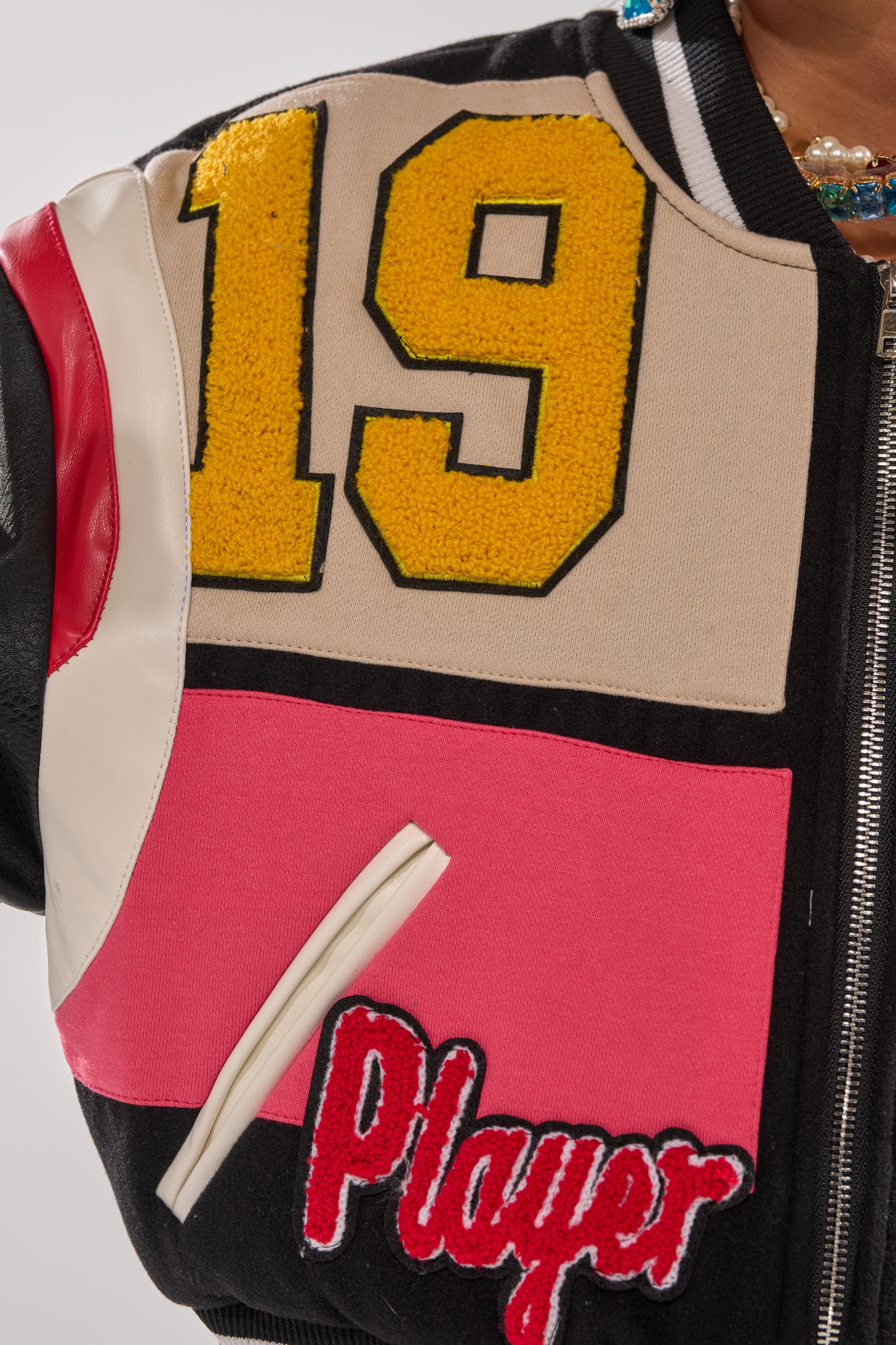 A close-up of the NEW YORK NEW YORK VARSITY BOMBER, featuring a large yellow "19," red and white panels, a zippered pocket, a red "Player" patch, and a partially visible necklace near the collar.