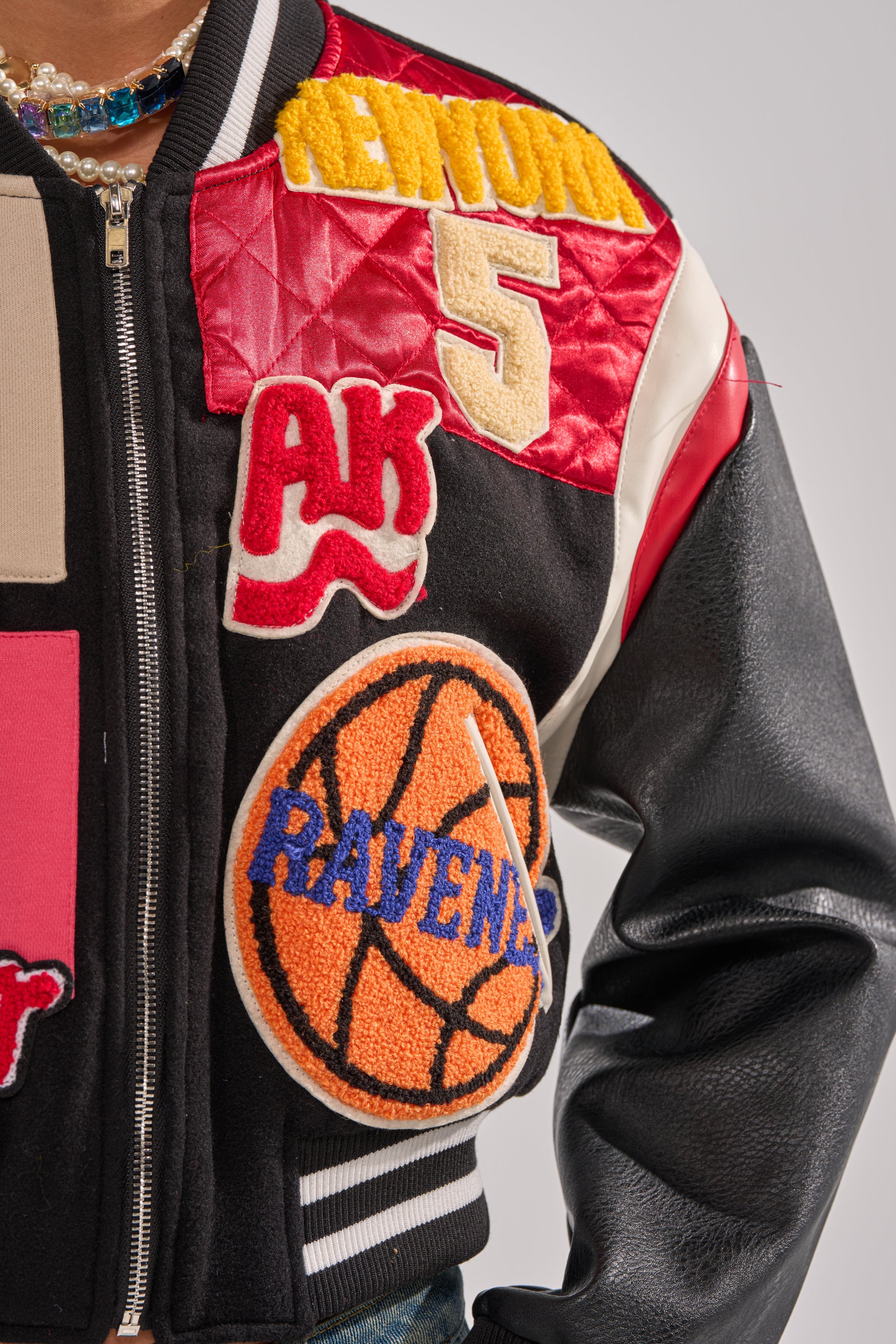 A close-up of the NEW YORK NEW YORK VARSITY BOMBER shows color-blocked fabric with varsity patches: a yellow "REMIND," red "AK," gold "5," and an orange basketball patch labeled "RAVENS.
