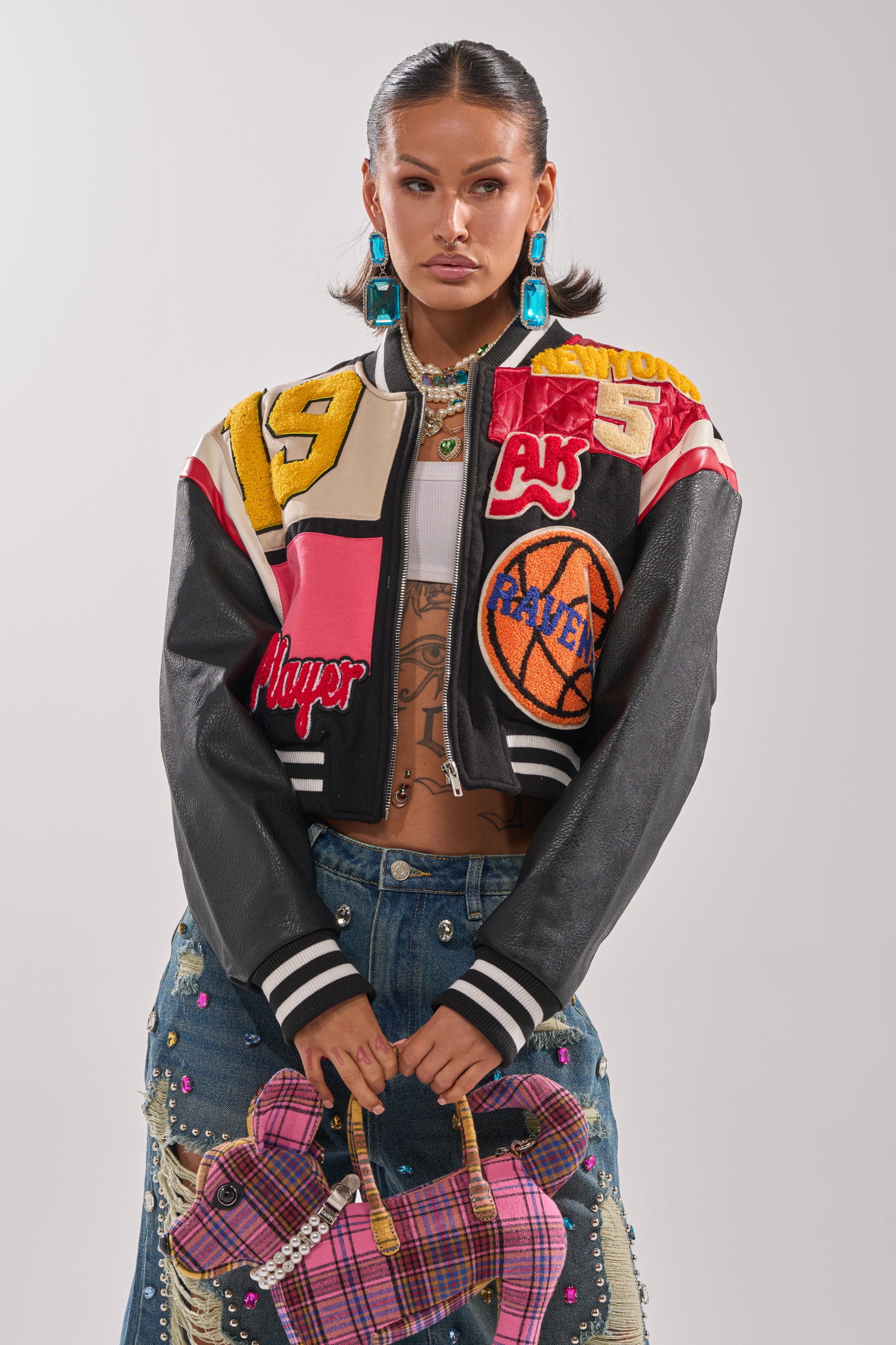 A woman wears the NEW YORK NEW YORK VARSITY BOMBER with basketball patches, a studded denim skirt, bold turquoise earrings, and holds a pink plaid dog-shaped handbag. Her sleek hair stands out against a plain white background.