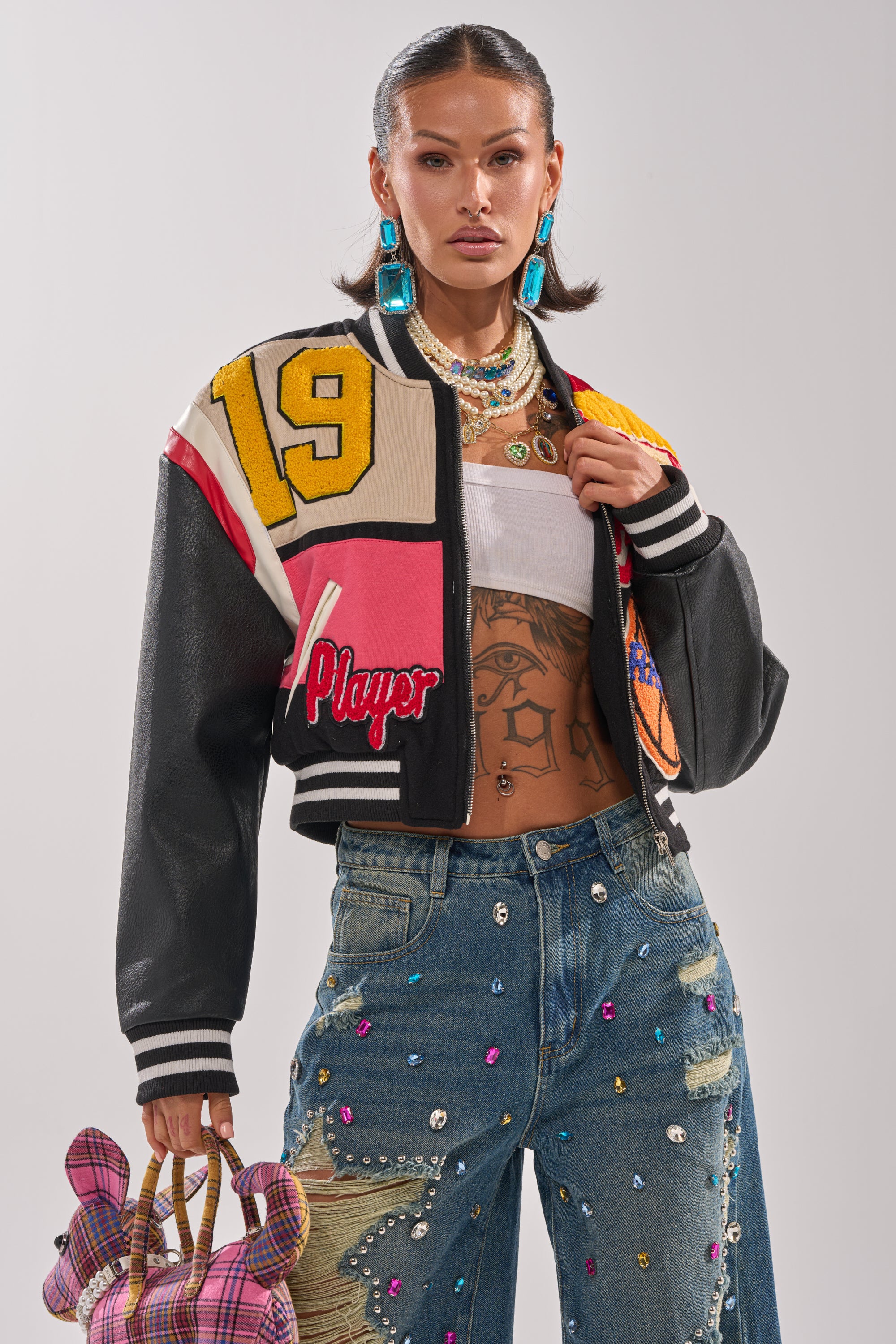 A stylish woman poses confidently in the NEW YORK NEW YORK VARSITY BOMBER jacket, jeweled jeans, blue earrings, layered necklaces, and a pink plaid teddy bear handbag. She sports slicked-back hair and bold makeup.