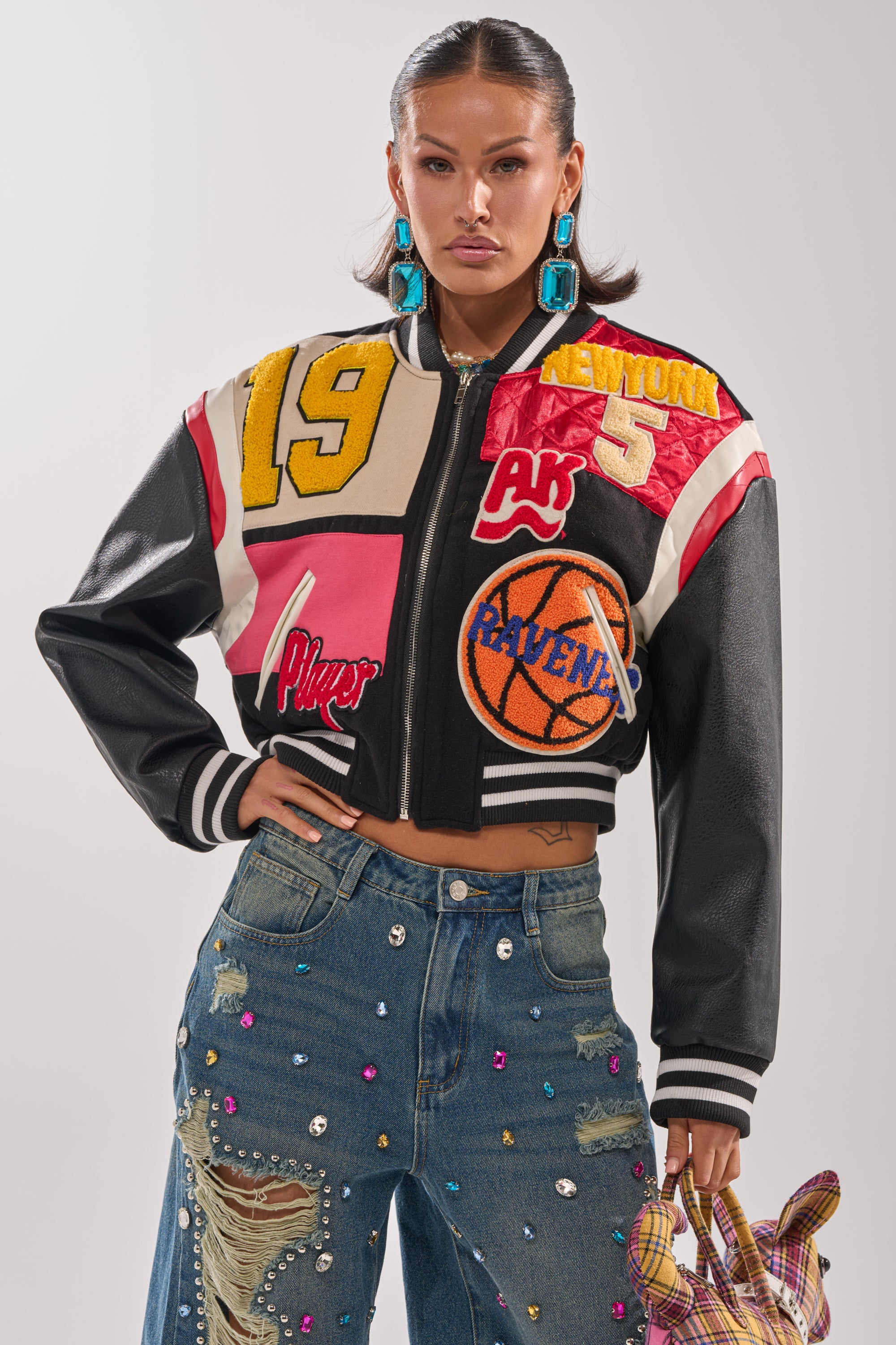 A woman models the NEW YORK NEW YORK VARSITY BOMBER with letterman patches, paired with rhinestone-studded distressed jeans, bold turquoise earrings, and a small plaid bag, posing confidently against a plain background.