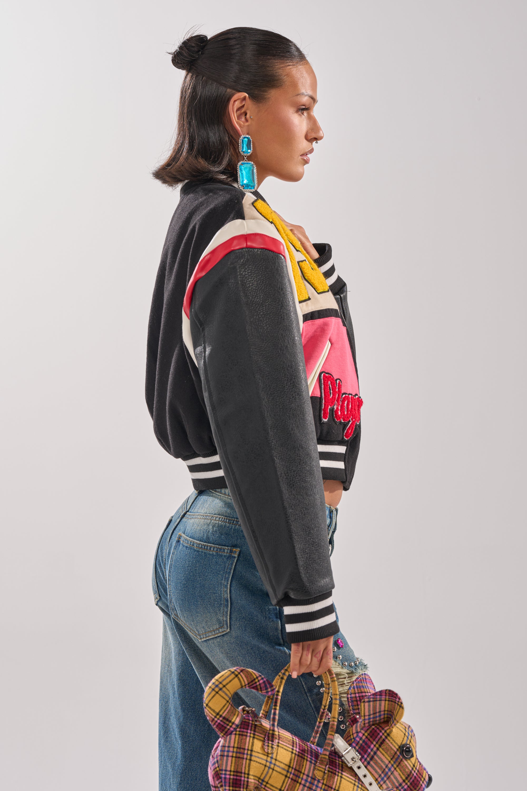A woman in a NEW YORK NEW YORK VARSITY BOMBER, blue jeans, and bold blue earrings holds a plaid teddy bear-shaped bag while standing in profile against a plain light background.
