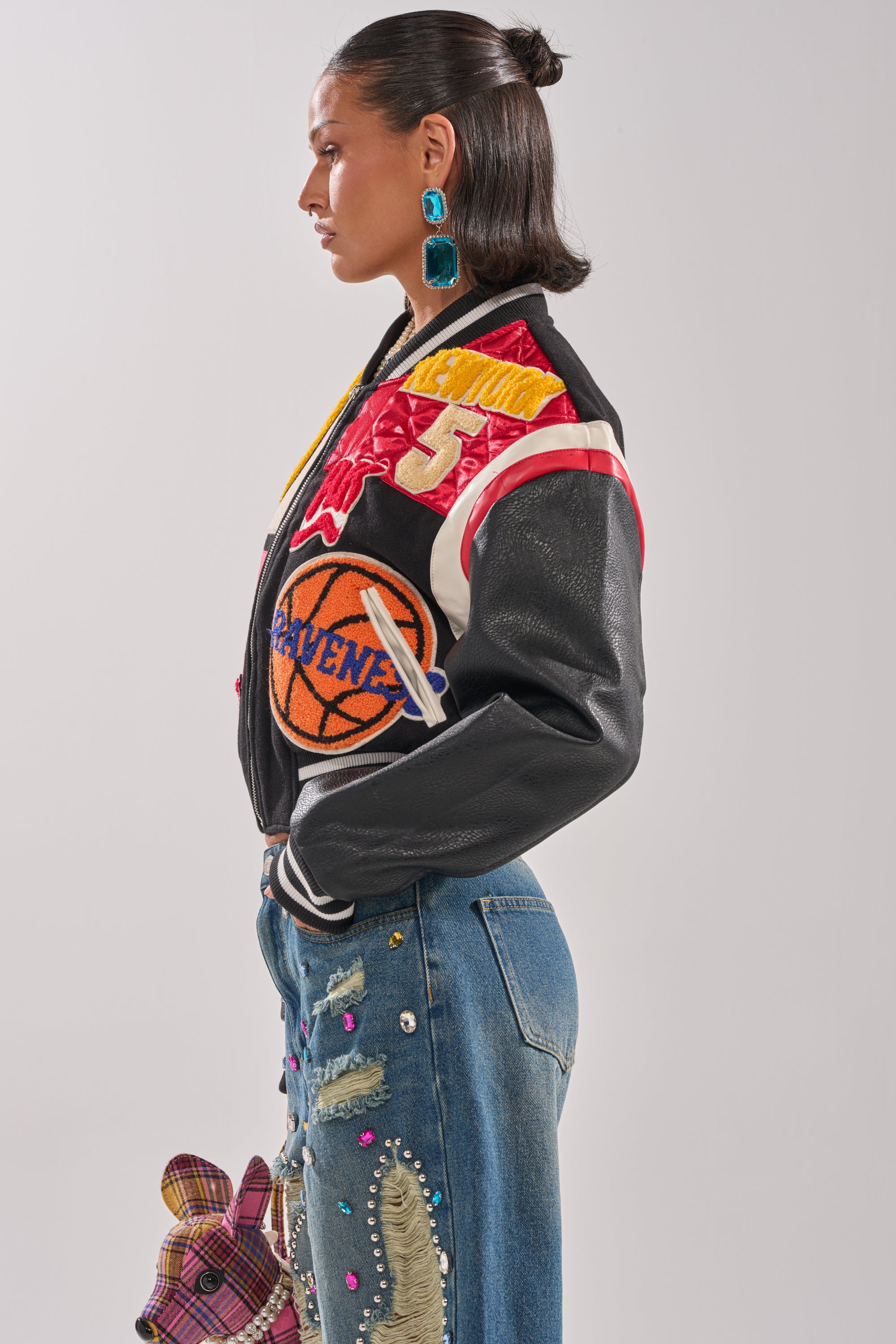 A woman in profile wears the NEW YORK NEW YORK VARSITY BOMBER—black with red, yellow, and orange patches—paired with blue statement earrings and embroidered blue jeans featuring colorful designs and a dog applique.