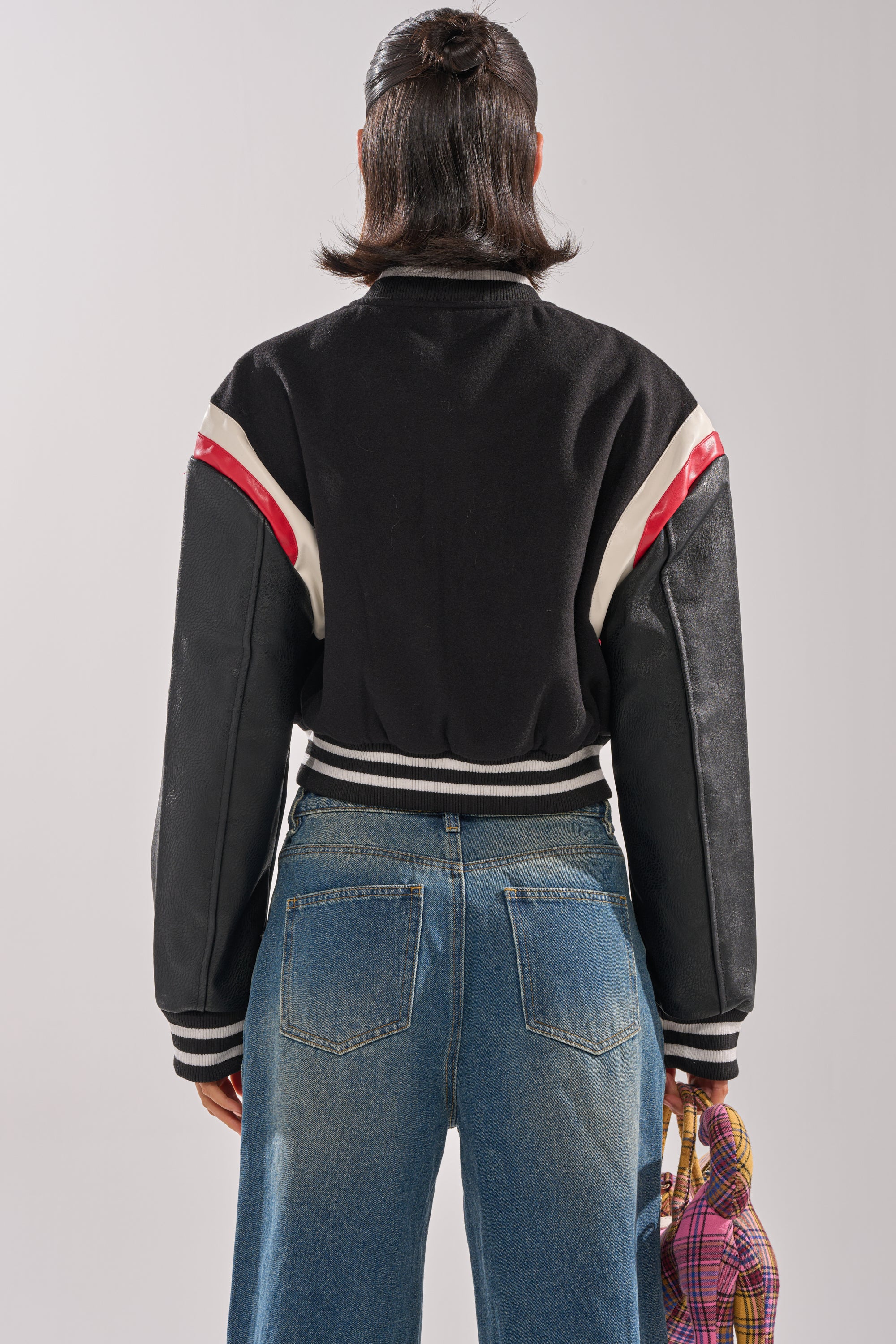 A person with shoulder-length dark hair, seen from behind in blue jeans and holding a small patterned handbag, wears the NEW YORK NEW YORK VARSITY BOMBER—a black jacket with red and white stripes.