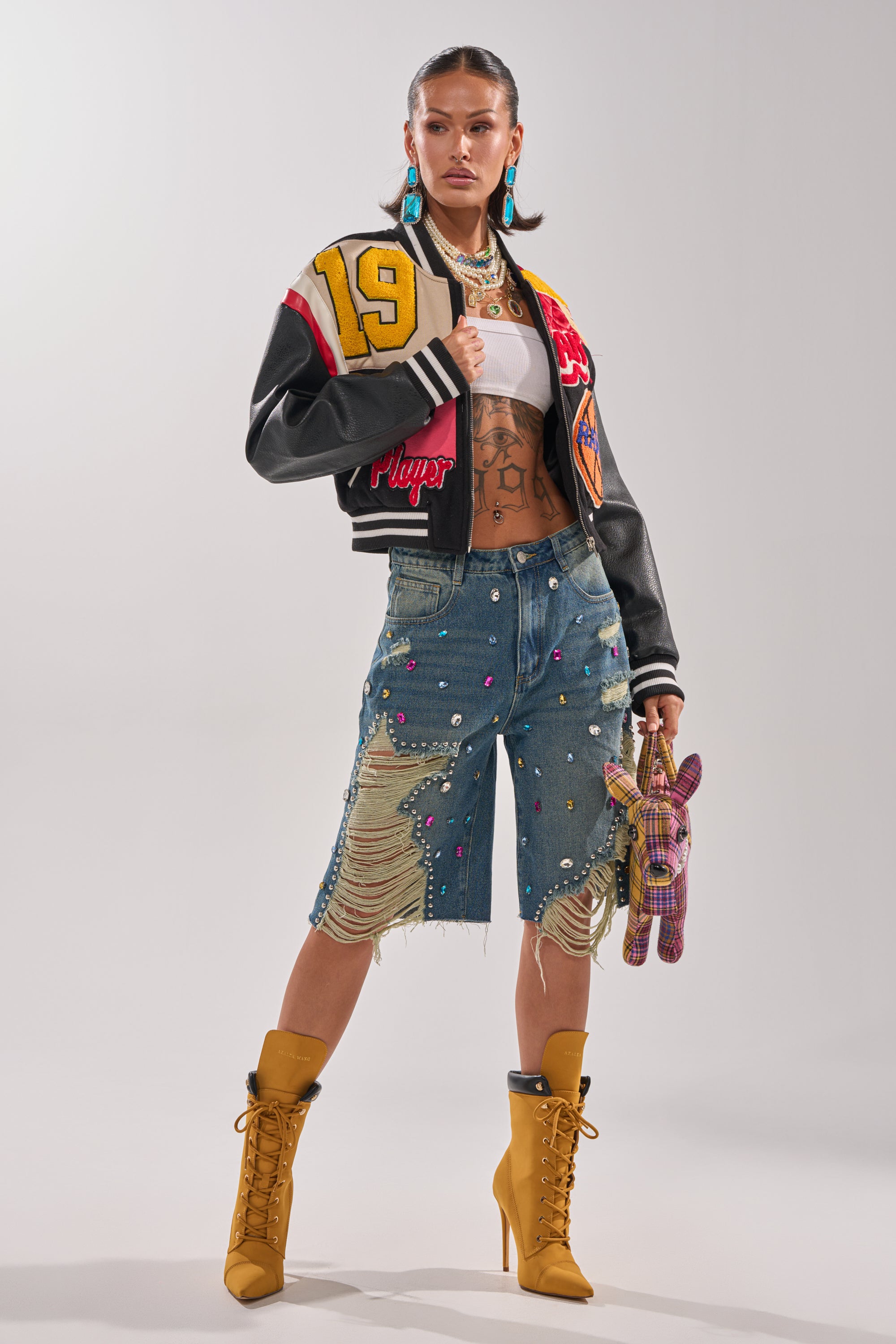 A woman poses confidently in the NEW YORK NEW YORK VARSITY BOMBER, paired with a white crop top, embellished ripped denim shorts, tan lace-up boots, and bold jewelry. She holds a colorful plaid stuffed animal and wears her hair sleek.