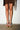 A person wearing a brown skirt stands against a white wall, showcasing their legs and feet in AZALEA WANG ARLETH GOLD SANDAL with chunky heels, pearl beads, gold chains, and turquoise accents.