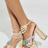 Close-up of someone wearing AZALEA WANG ARLETH GOLD SANDAL with gold platform and chunky block heels, adorned with white bead straps and colorful shell embellishments, elegantly positioned against a neutral background.