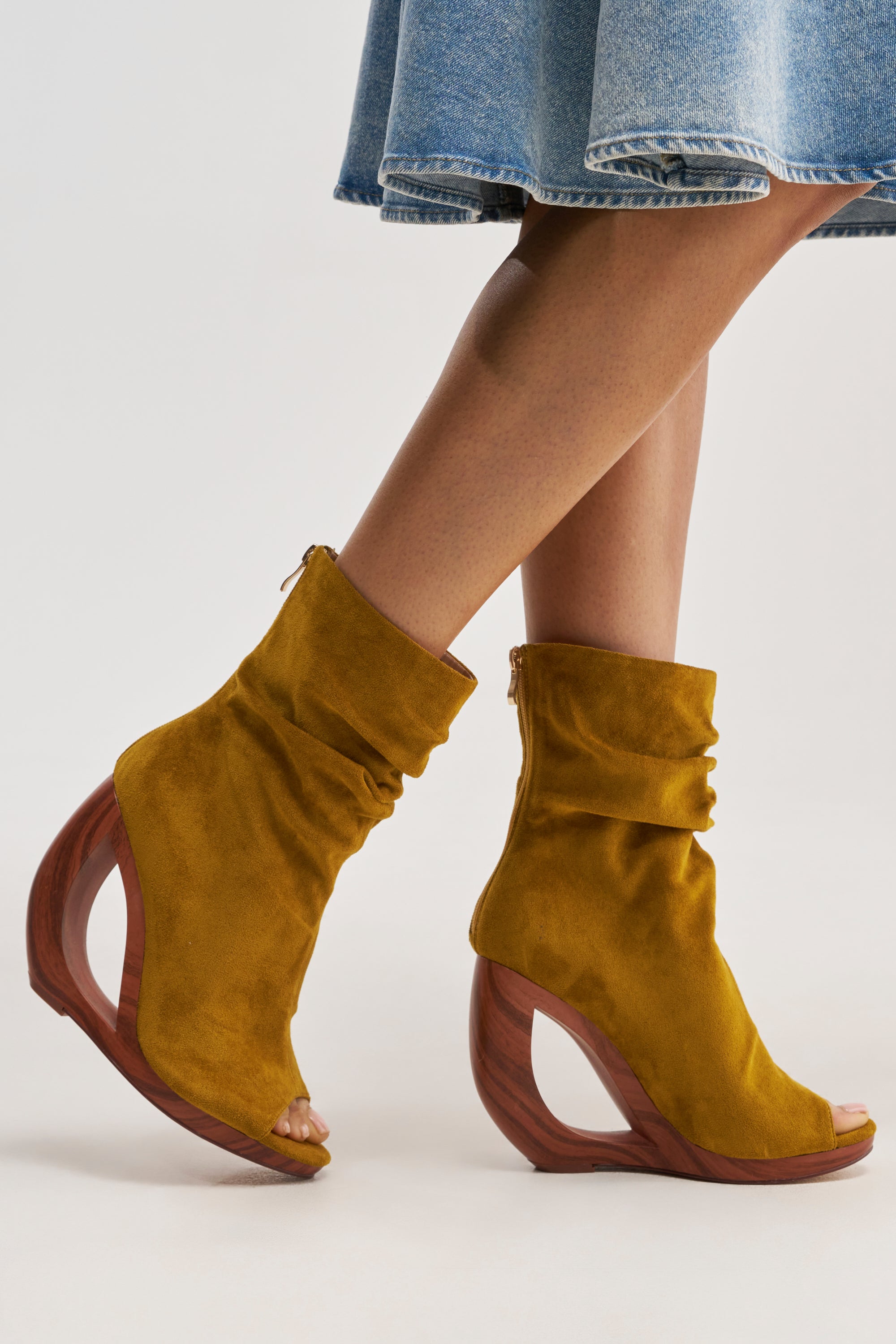 A person wears a denim skirt and the AZALEA WANG ARLO Mustard Wedge Open Toe Faux Suede Bootie, featuring curved wooden wedge heels, standing on a light background.