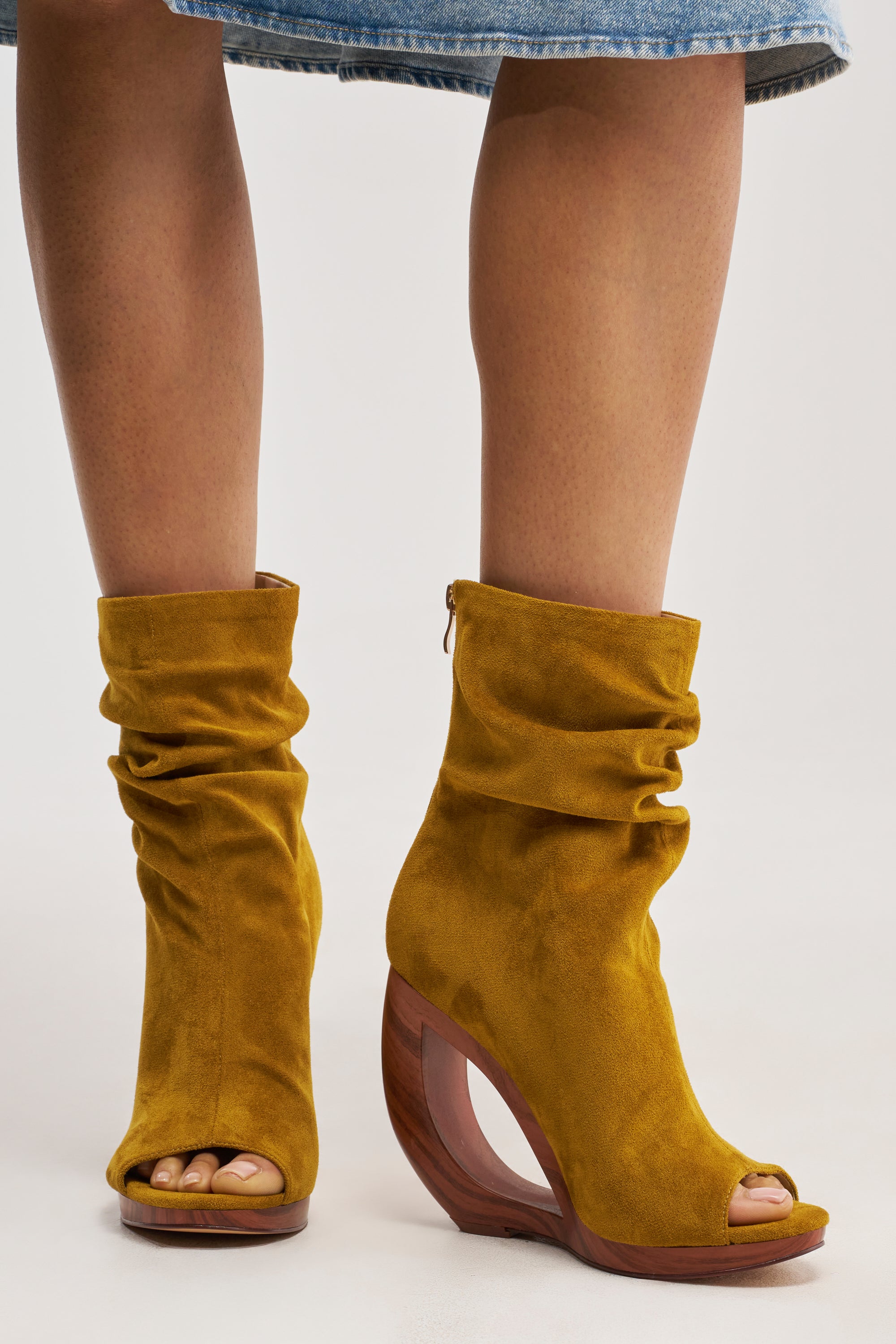 A person wears the AZALEA WANG ARLO MUSTARD WEDGE OPEN TOE FAUX SUEDE BOOTIE, featuring a comfort cushion insole, slouchy suede design, and curved wooden heel, paired with a blue denim skirt.