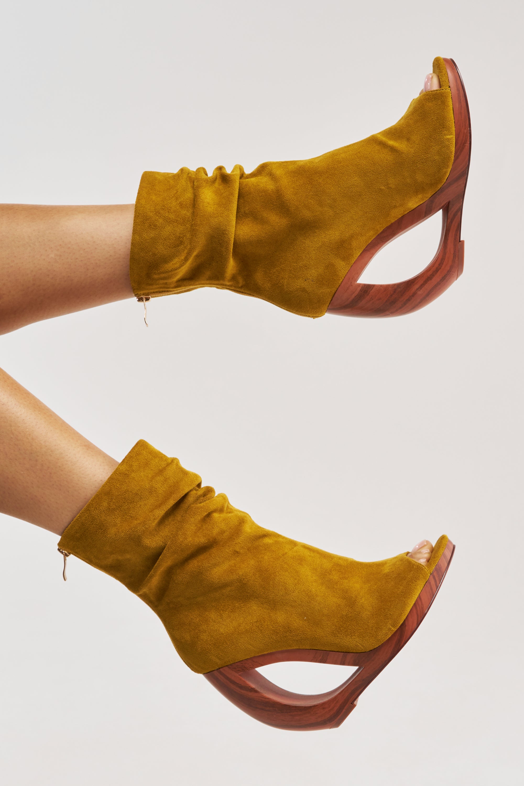 The AZALEA WANG ARLO Mustard Wedge Open Toe Faux Suede Booties feature chic curved wooden wedge heels and a comfort cushion insole, shown on crossed legs against a plain light background.