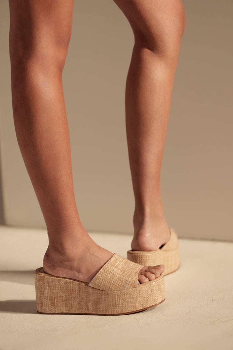 A person stands on a beige surface, wearing the AZALEA WANG ARROW RAFFIA WEDGE SANDAL with light tan raffia material, thick chunky wedge heels, and an open square toe. Their feet and lower legs are shown against a neutral background.