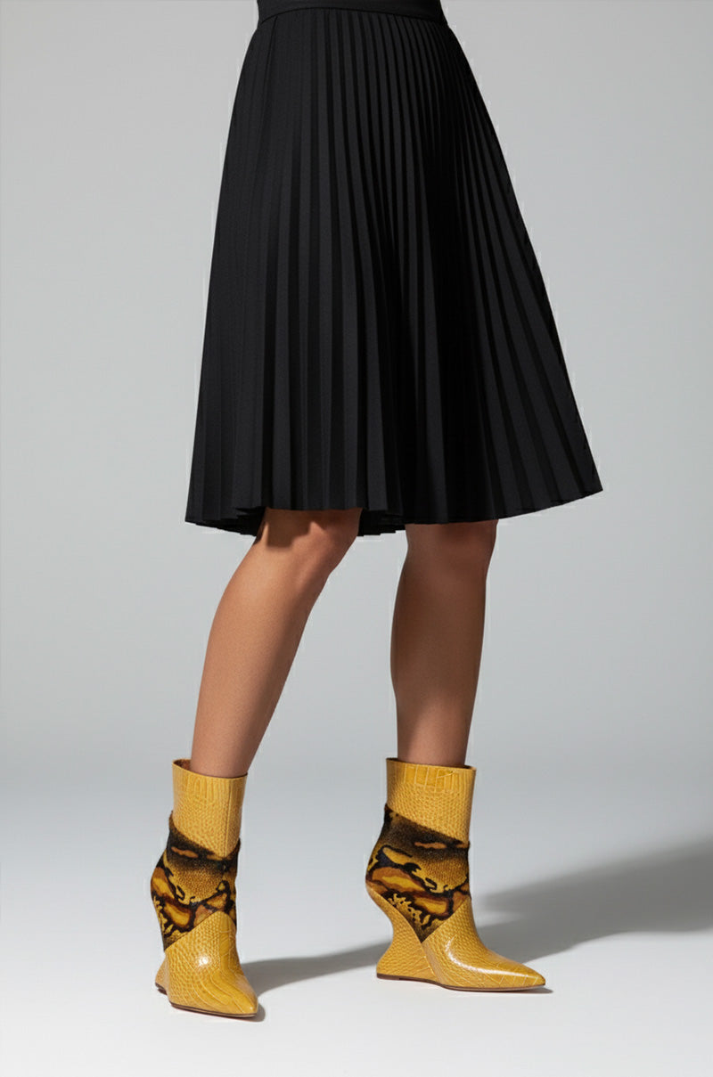 A person in a knee-length black pleated skirt and AZALEA WANG ARTOUL YELLOW COLOR BLOCKED YELLOW WEDGE BOOTIE with a black abstract pattern stands against a light gray background.