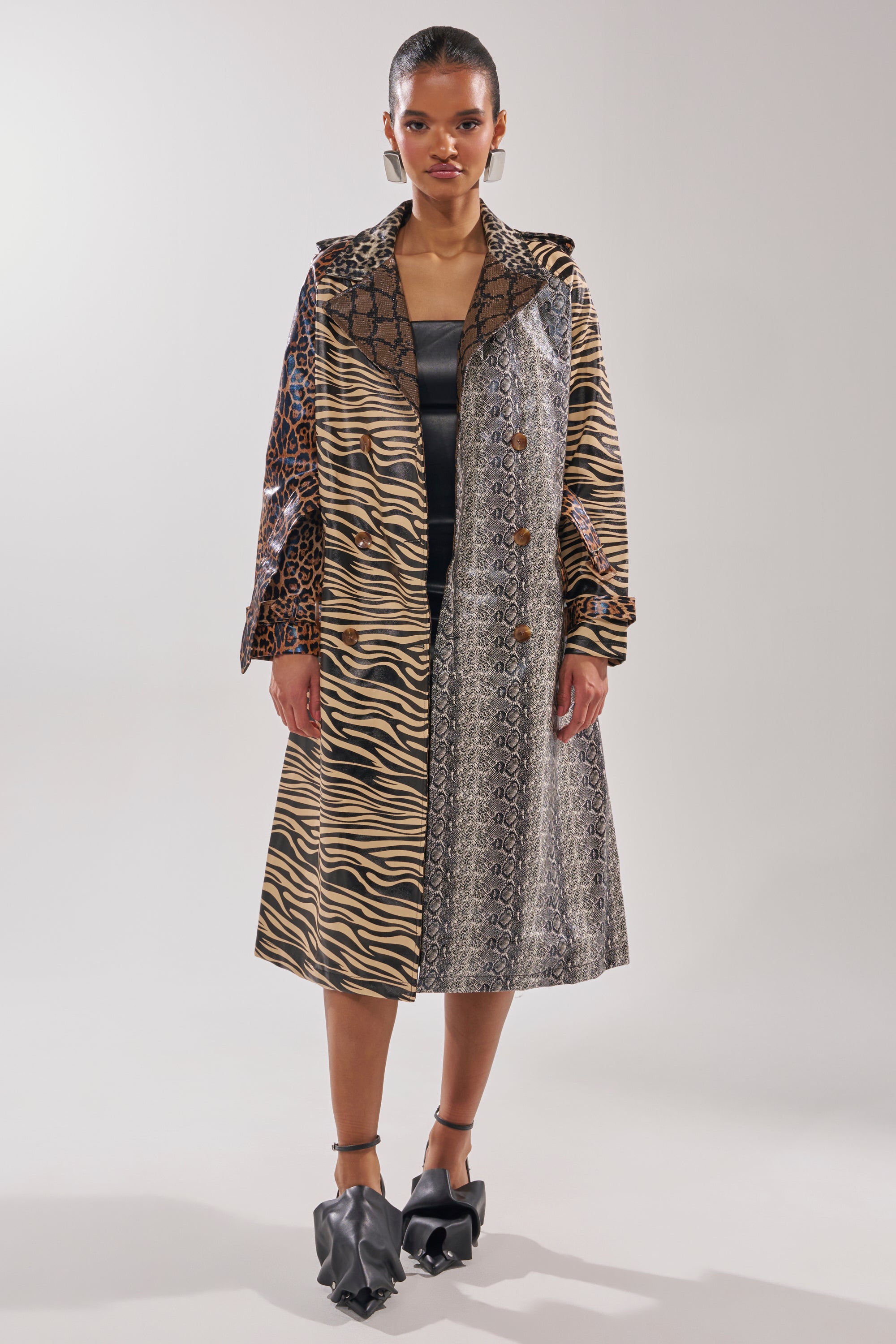 A woman wears the BASEL PAINTED TRENCH with animal prints and geometric designs, styled over a black dress and statement black ruffled shoes. She poses against a plain, light background.