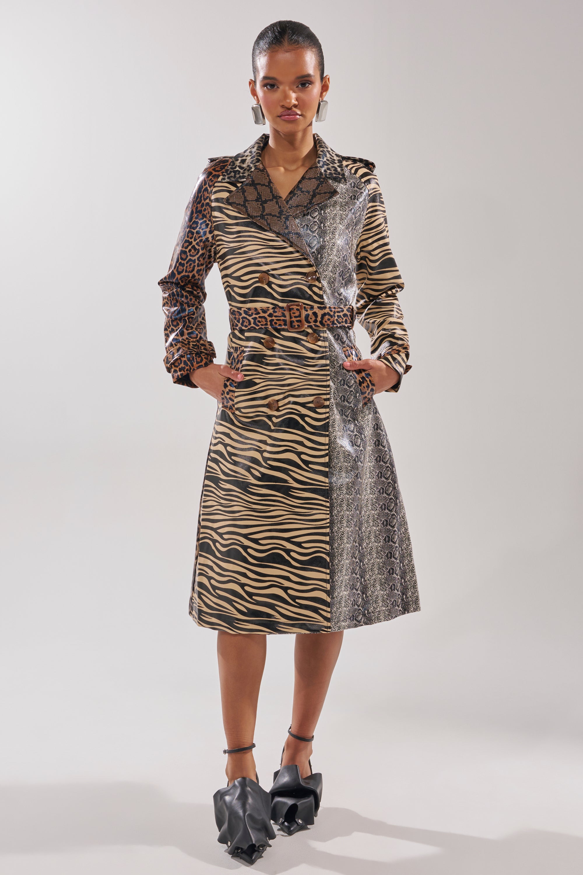 A woman stands facing forward in the BASEL PAINTED TRENCH, a longline animal print coat with zebra, leopard, and snake patterns. Her hands are in her pockets and she wears black pointed shoes with sculptural details.