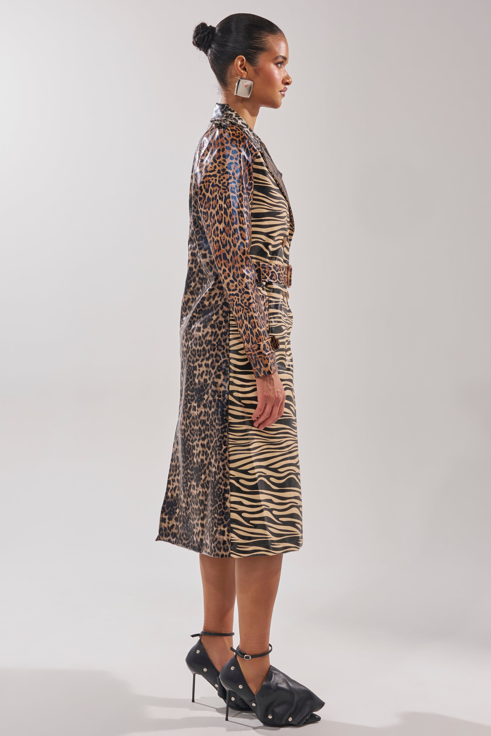 A woman in profile wears the BASEL PAINTED TRENCH, featuring leopard print on the back and sleeves with zebra print on the front. Her hair is in a bun, complemented by large earrings and black bow-adorned heeled shoes.