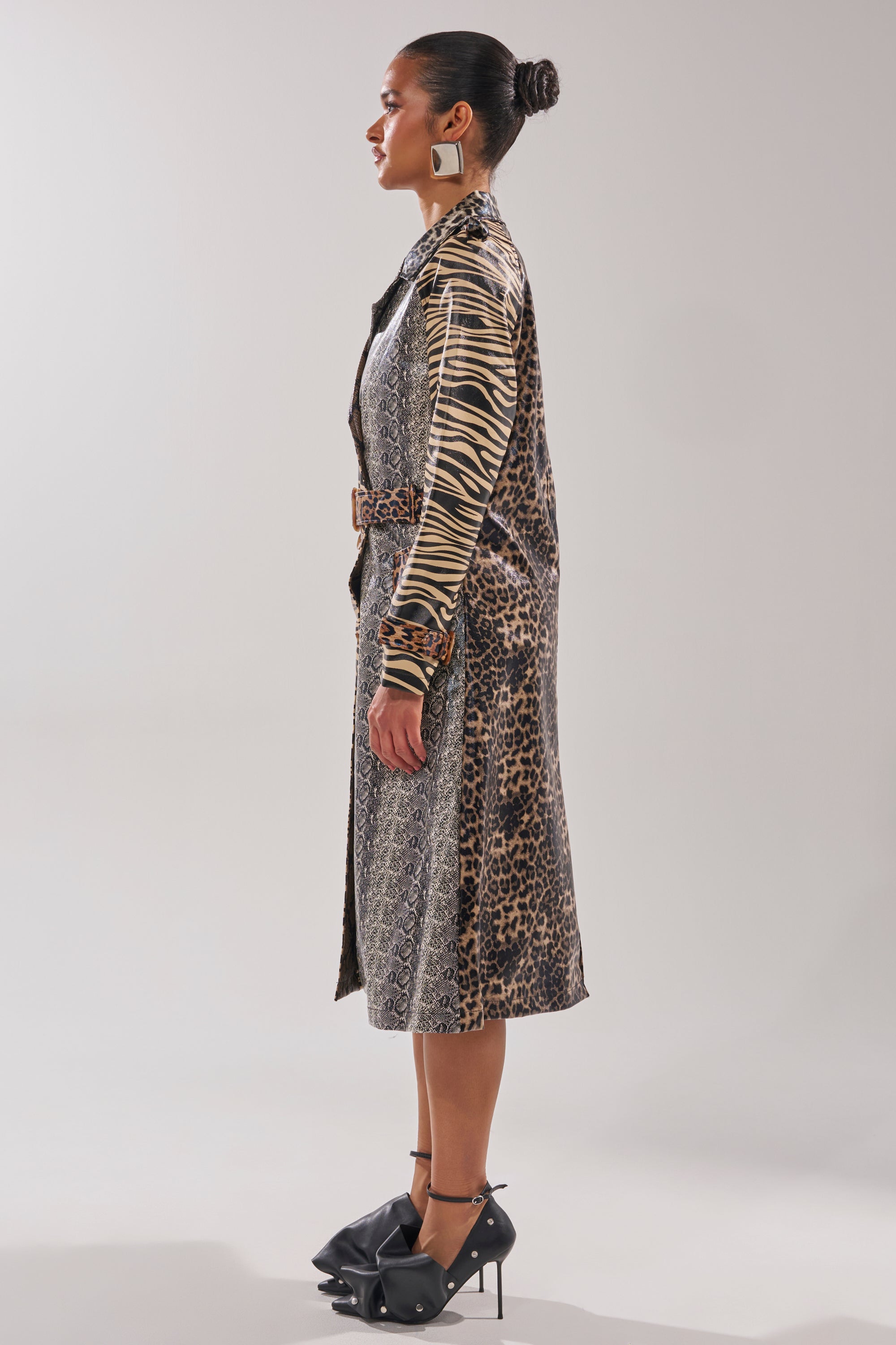 A woman in profile wears the BASEL PAINTED TRENCH, a longline animal print jacket, styled with statement earrings and black high heels with large bows, standing against a plain light background.