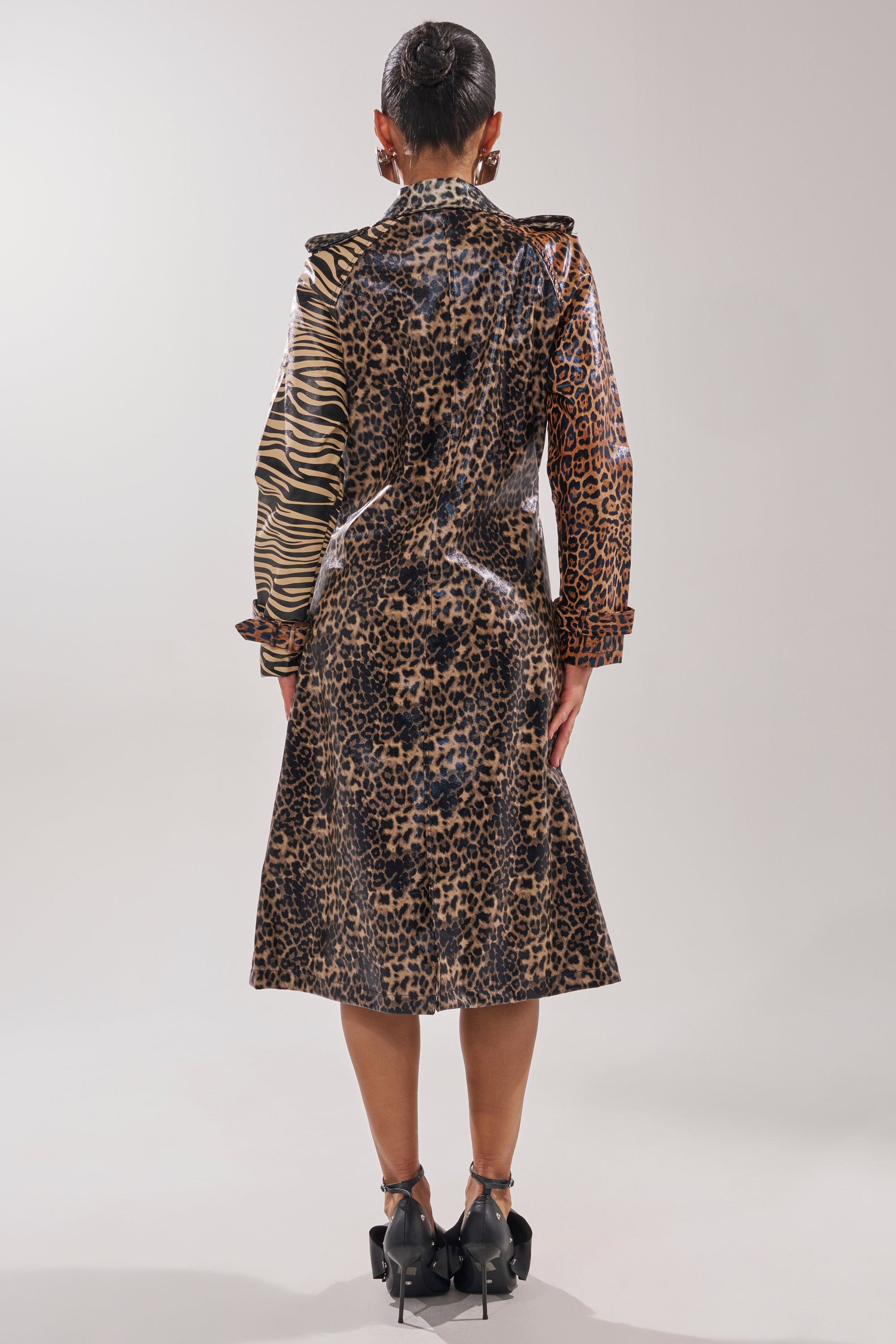 A person stands facing away, wearing the BASEL PAINTED TRENCH—a long, glossy coat with mixed leopard and zebra prints. Their hair is in a bun, paired with black heels against a plain, light background.