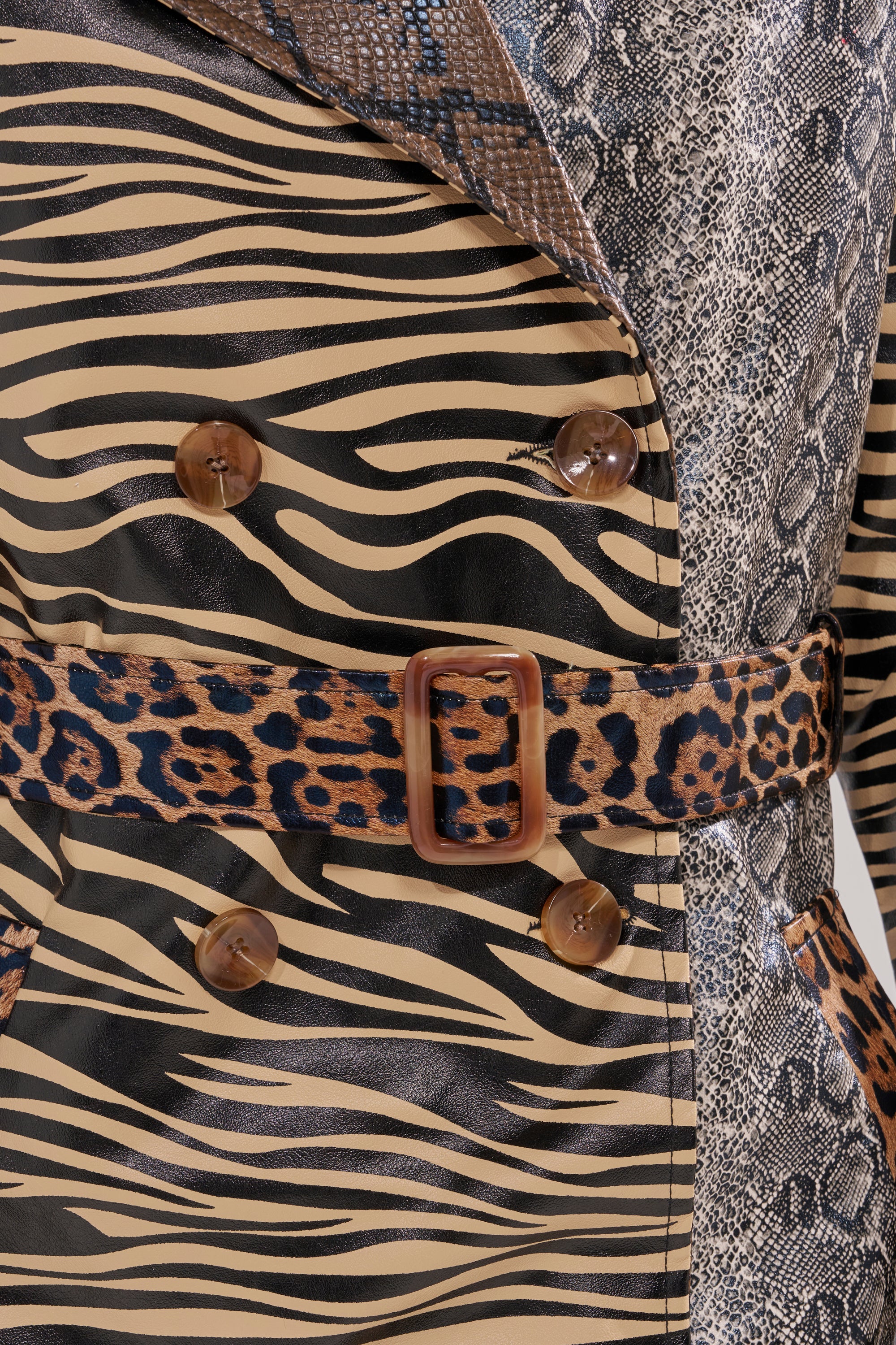 Close-up of the BASEL PAINTED TRENCH, a striking longline jacket with animal prints—zebra stripes, snakeskin, and leopard spots—brown buttons, and a leopard-print belt with a wooden buckle.