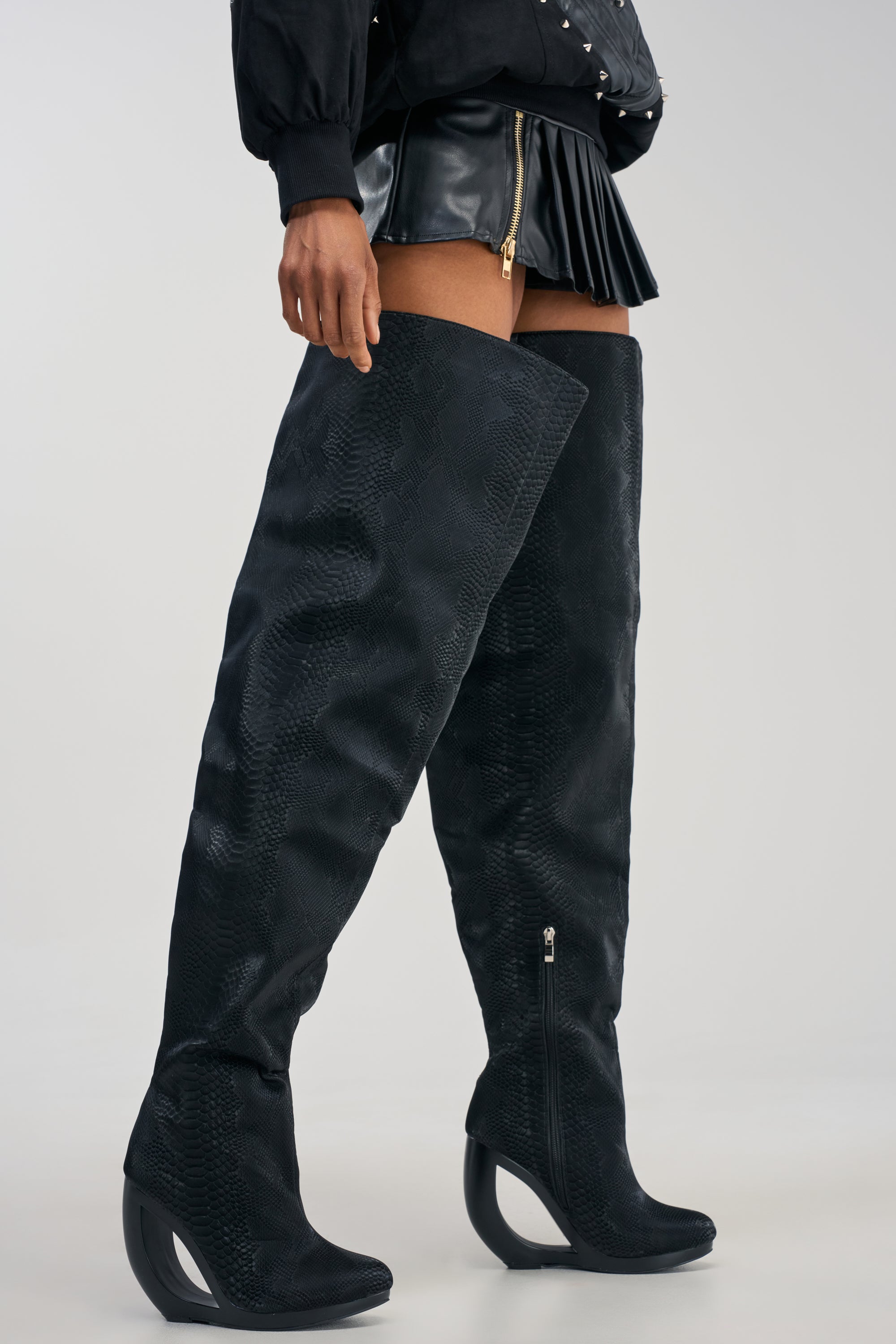 A person wearing a black pleated mini skirt and the AZALEA WANG ASA BLACK SNAKE OPEN WEDGE THIGH HIGH BOOT, featuring striking curved heels, stands on a light background.