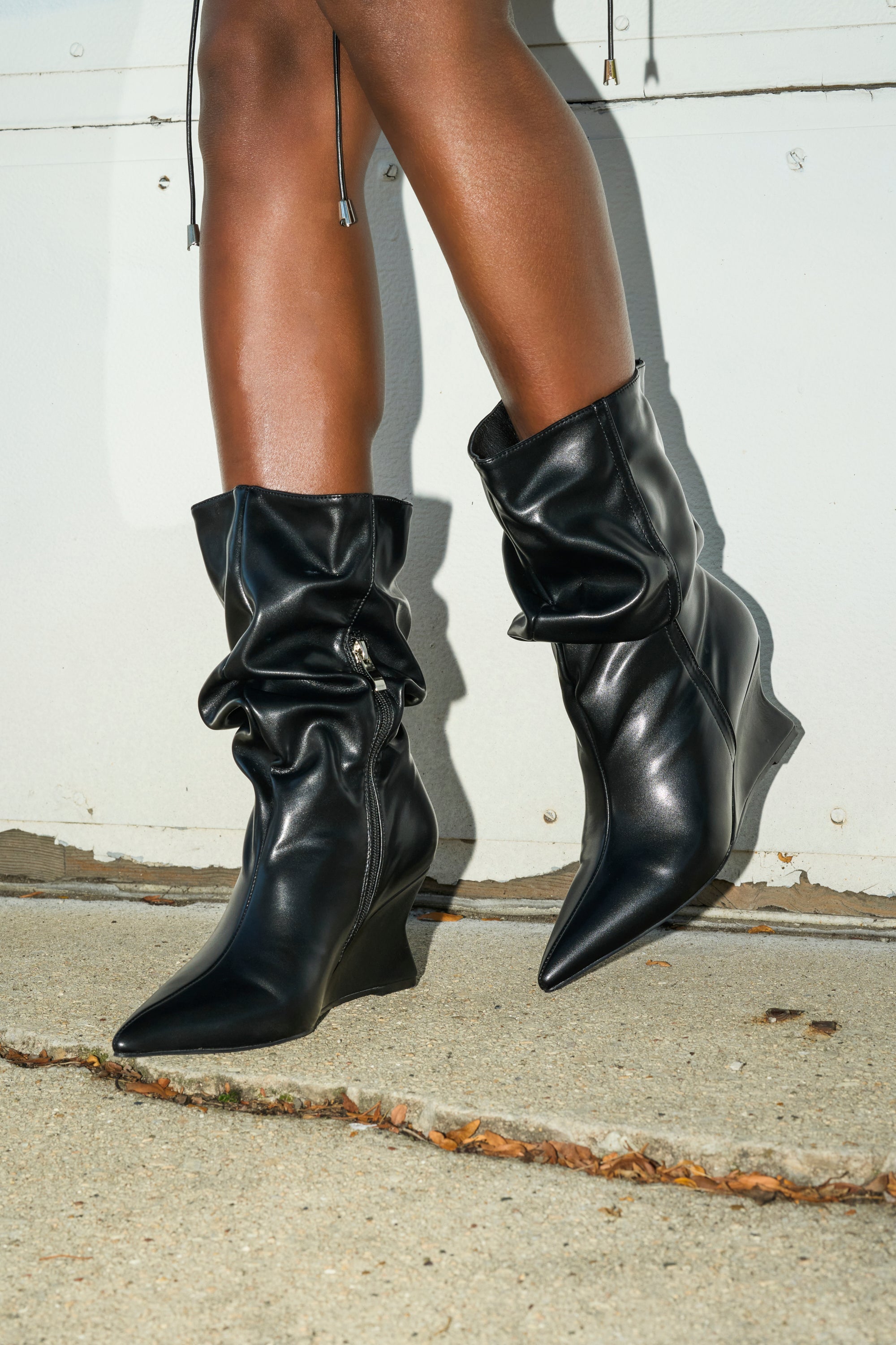 A person models the AZALEA WANG ASAF RUCHED WEDGE BOOTIE IN BLACK, standing on concrete before a light wall, with only their lower legs and feet in view.