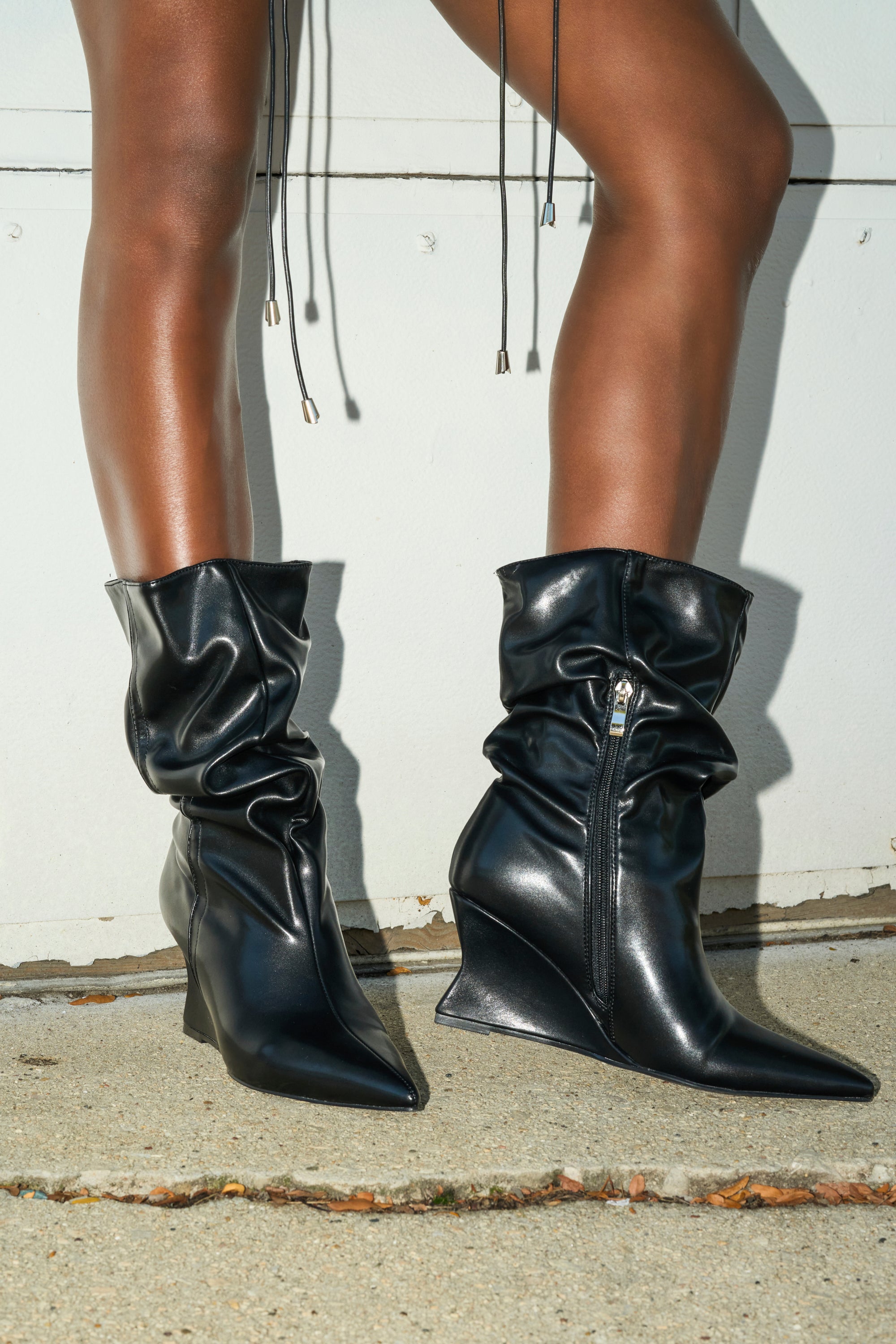 Someone stands on concrete in front of a white wall, wearing the AZALEA WANG ASAF RUCHED WEDGE BOOTIE IN BLACK. These shiny faux leather booties feature pointed toes, side zippers, and gathered ruched details for a modern look.