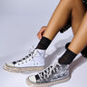 A person wearing the ASH BLACK WHITE EMBELLISHED SNEAKER with mismatched styles adorned with rhinestone shoelaces. The left sneaker is plain white, while the right features a striking black and white sequin pattern. Both sneakers have studded soles with metallic spikes, paired perfectly with sheer black ankle socks.