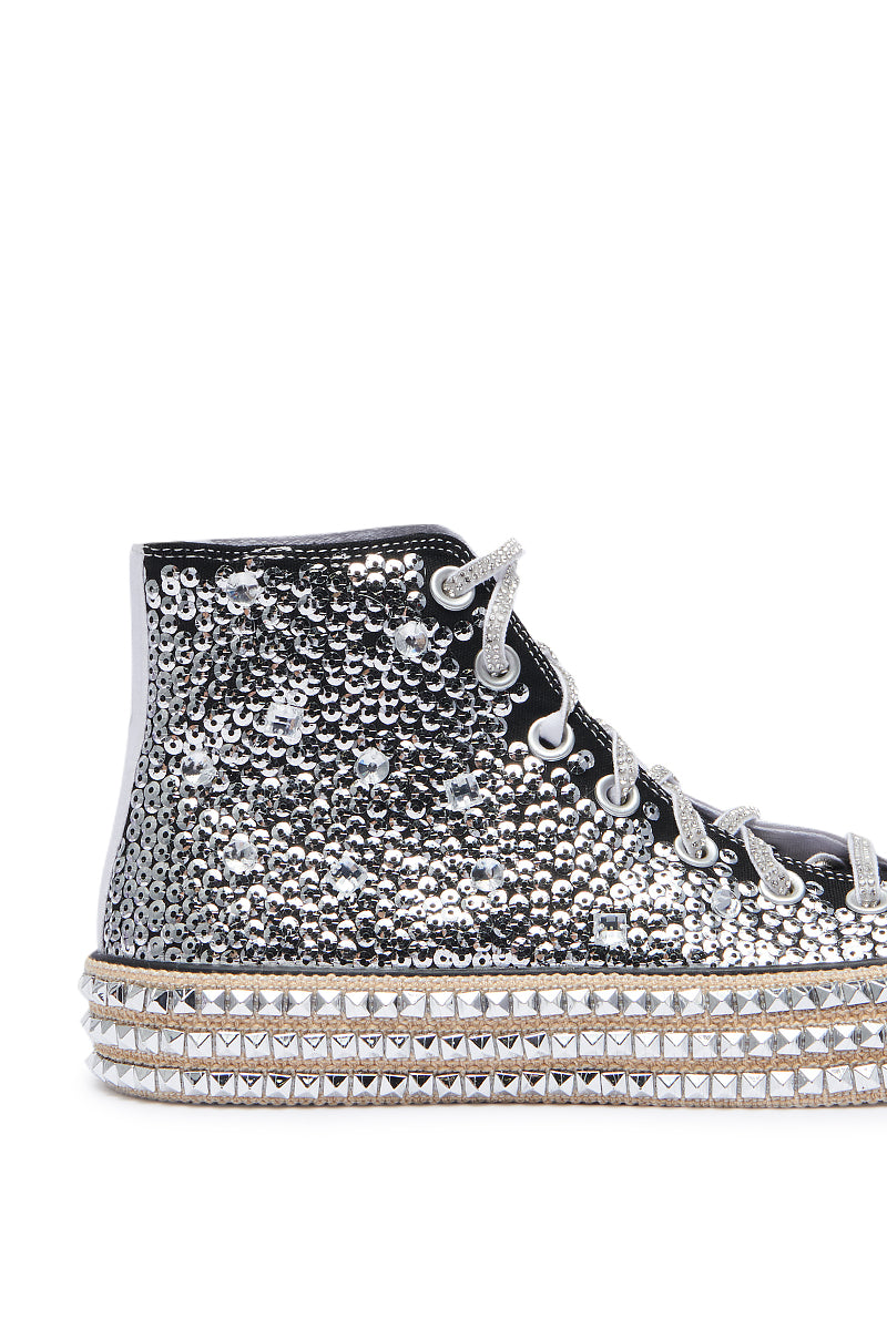 The ASH BLACK WHITE EMBELLISHED SNEAKER is a high-top shoe covered in silver sequins and adorned with sparkling rhinestones. It features shoelaces embellished with rhinestones and a thick platform sole accented with silver studs, resulting in an overall dazzling, eye-catching design.
