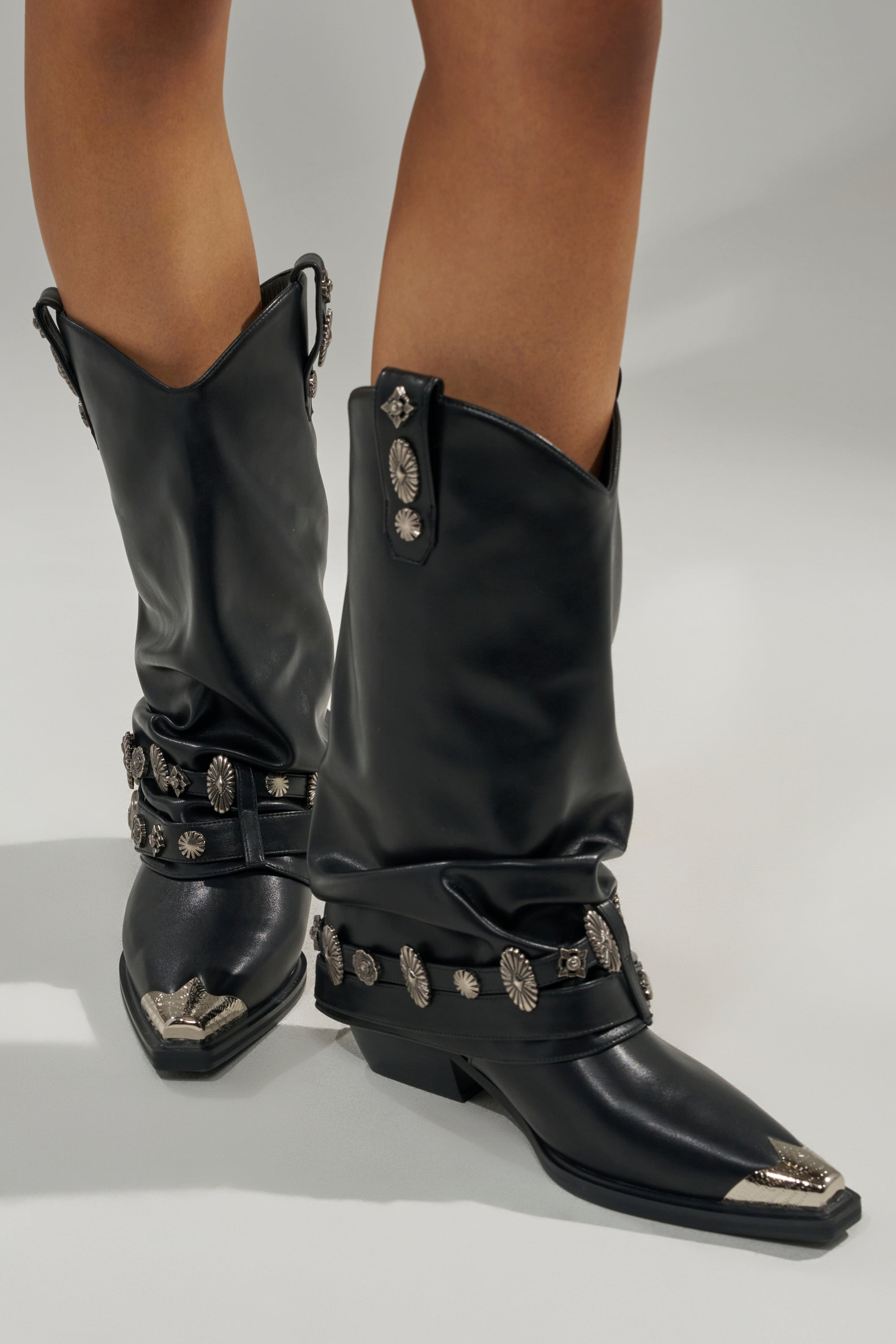 Someone wearing the AZALEA WANG ASHRAF BLACK PU SILVER HARDWARE WESTERN BOOTIE—with silver hardware, decorative conchos, chunky heel, and pointed metal-tipped toes—stands on a light-colored surface.