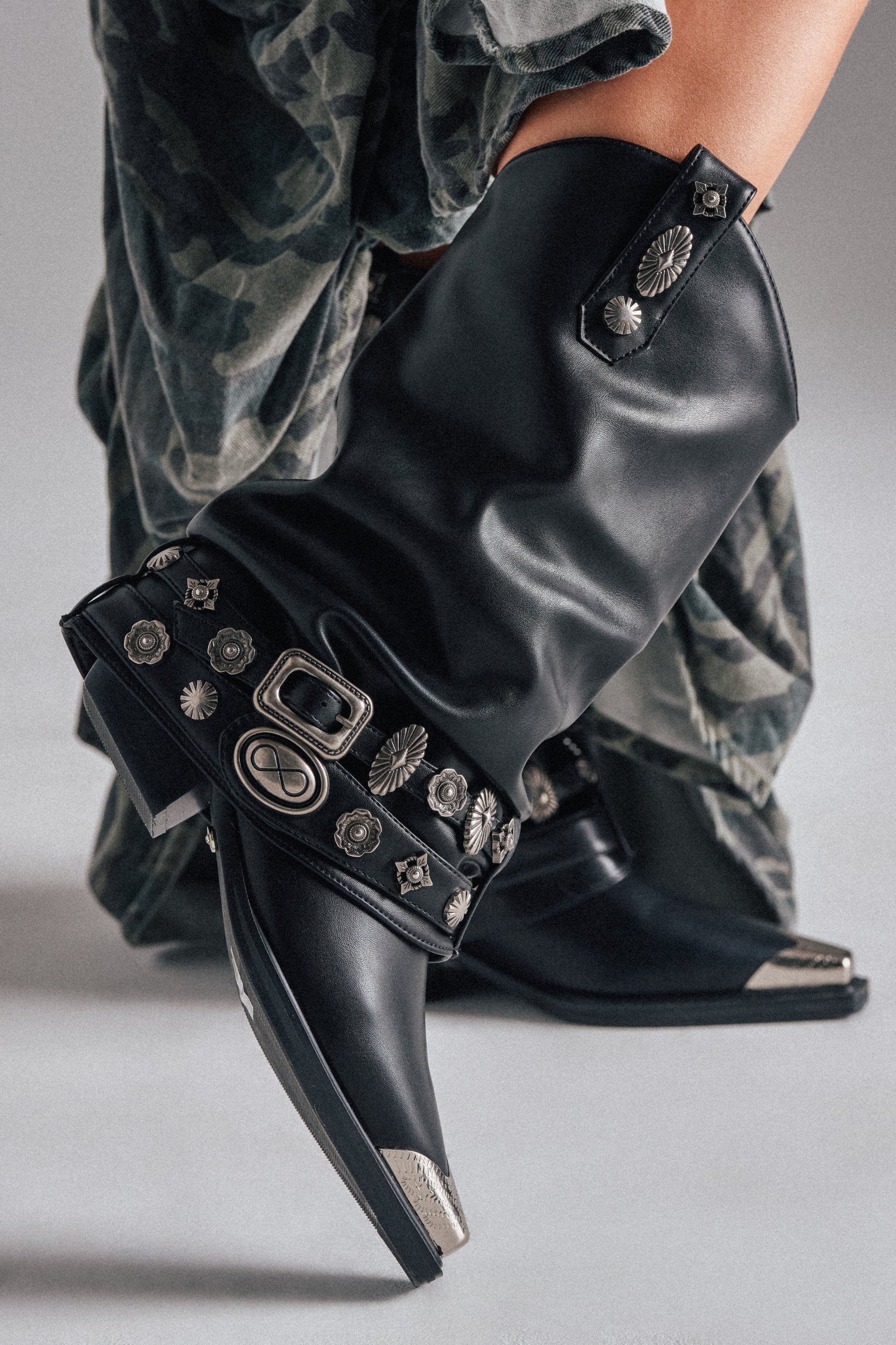 A close-up of a person wearing the AZALEA WANG ASHRAF BLACK PU SILVER HARDWARE WESTERN BOOTIE with silver buckles and studs, paired with a camouflage skirt.