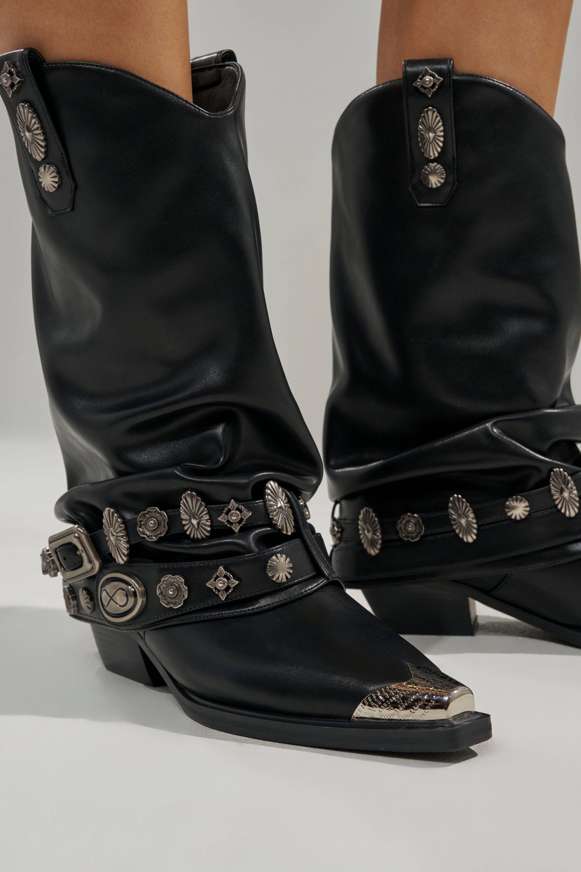 The AZALEA WANG ASHRAF Black PU Silver Hardware Western Bootie features black faux leather, silver conchos, buckles, detailed straps, a pointed toe with metal cap, and a chunky heel. Photographed against a light background.