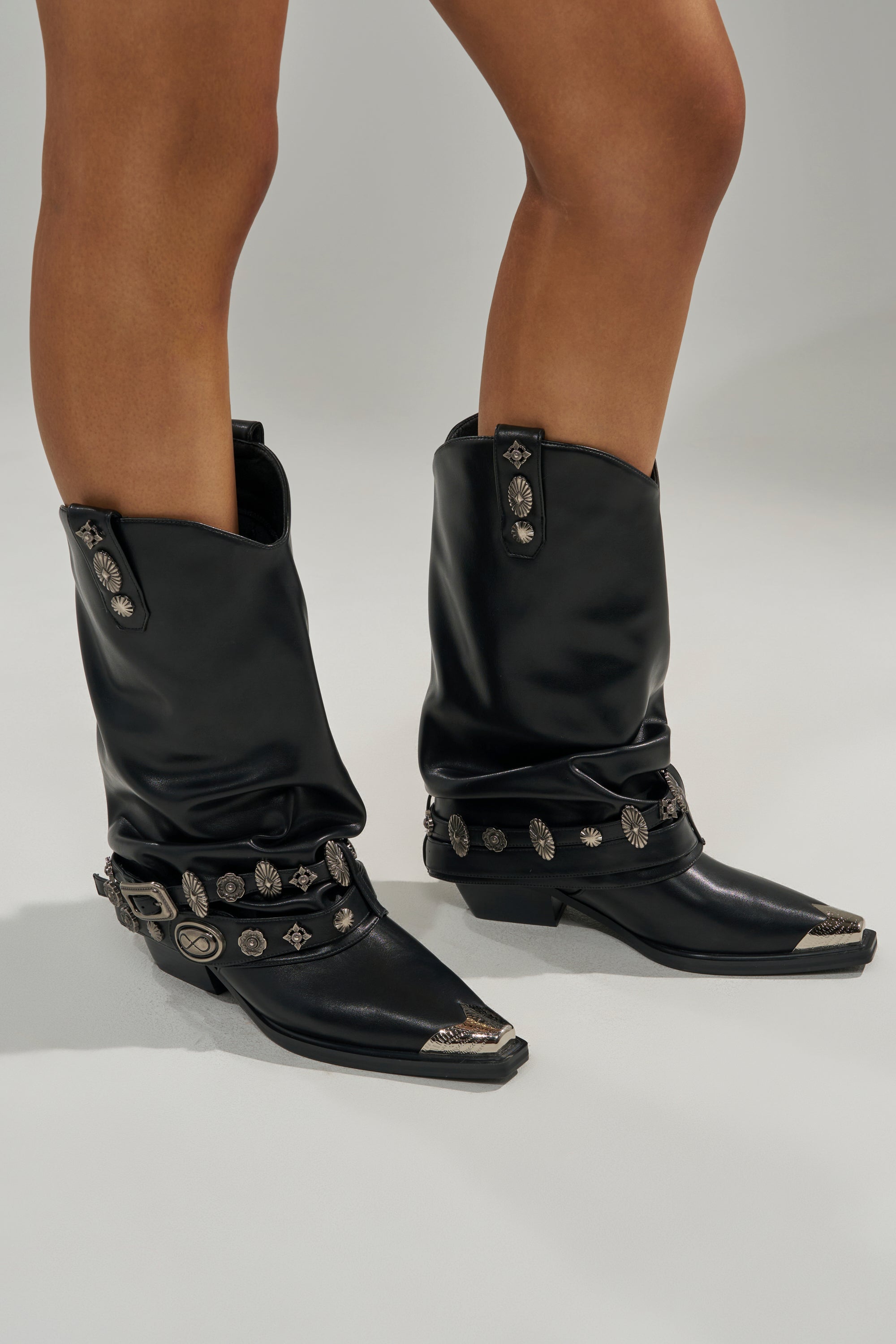 A person stands on a neutral background wearing the AZALEA WANG ASHRAF Black PU Silver Hardware Western Bootie, featuring pointed toes, silver embellishments, and a chunky heel.