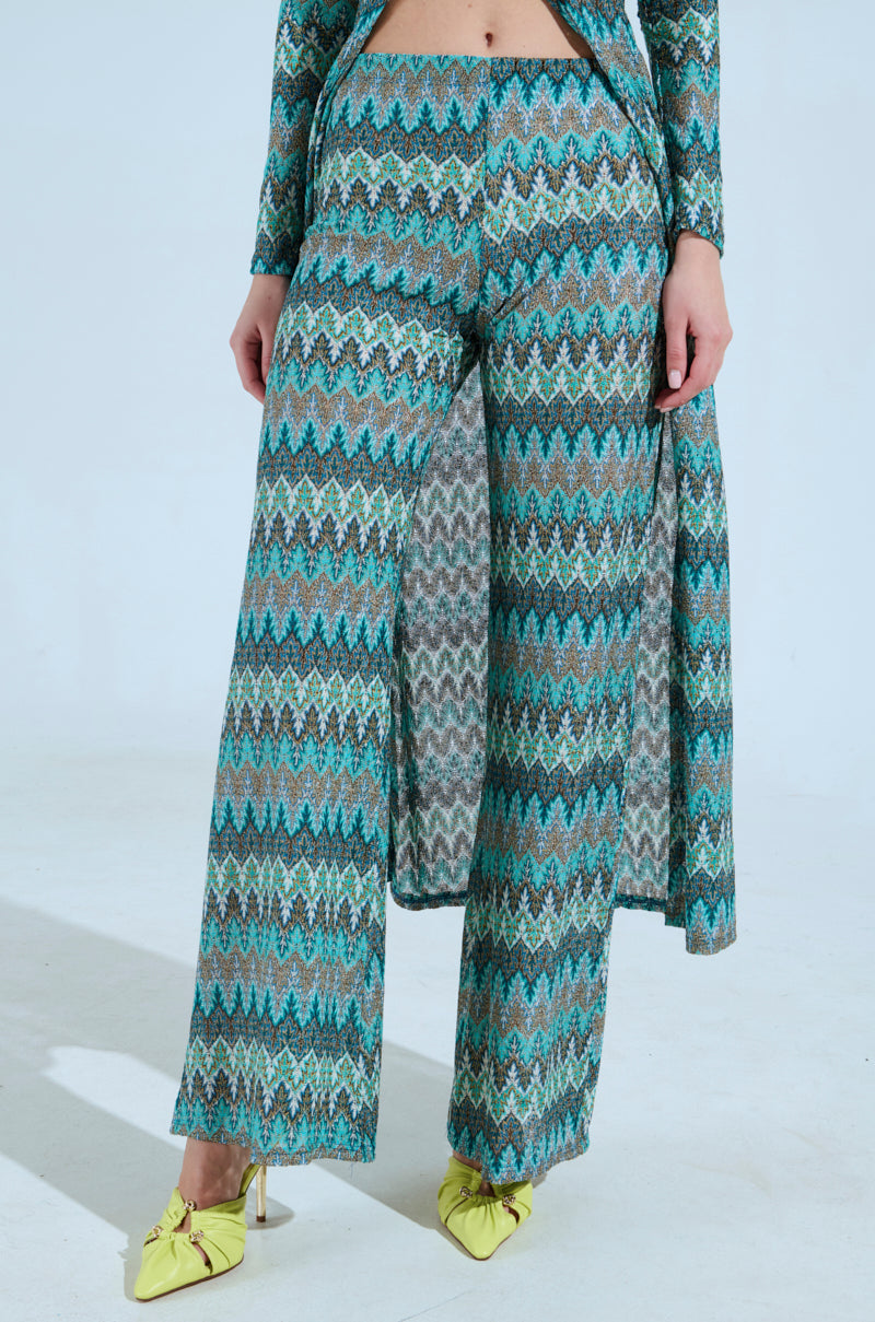 A person is wearing SHOW ME SOMETHING PANT, a patterned outfit with shades of blue, green, and black in a chevron design. The high-rise fit ‘n flare pants are wide-legged and paired with bright yellow pointed-toe shoes against a plain light background. 0