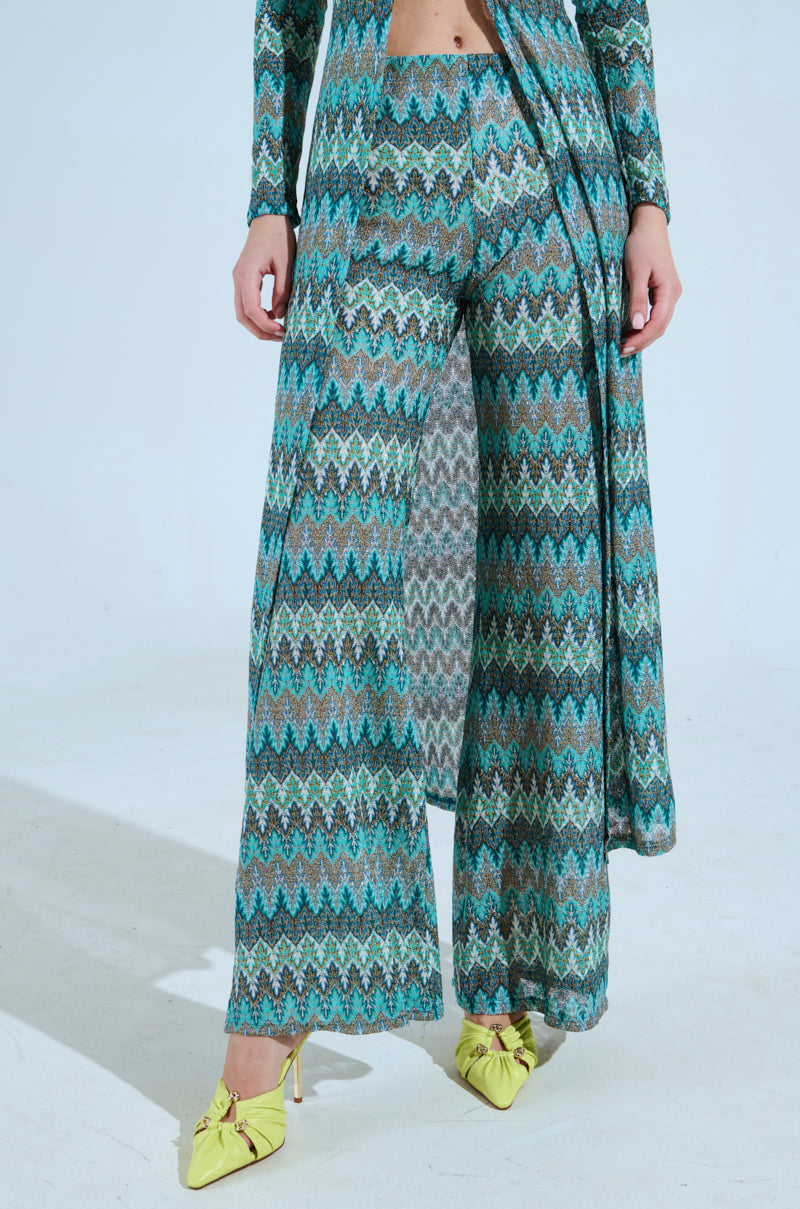 A person wears the SHOW ME SOMETHING PANT, a long, patterned outfit with a chevron design in green, blue, and brown. It features a stretchy knit top with a split front and high-rise fit 'n flare wide-legged pants. Yellow high-heeled shoes with bows complete the look. 1