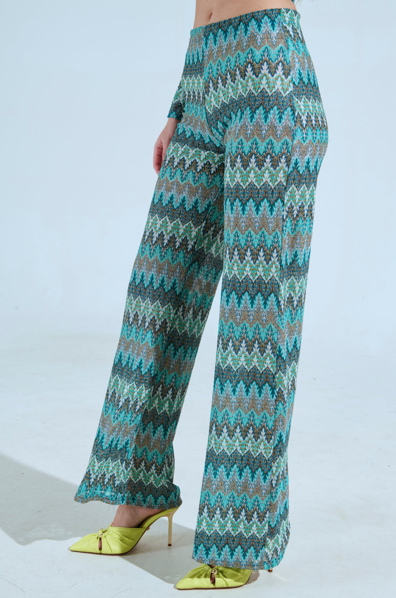 A person stands against a plain backdrop wearing SHOW ME SOMETHING PANT, featuring high-waisted, wide-leg design with a stretchy knit fabric in vibrant blue, green, and beige chevron pattern. They pair them with lime green pointed-toe heels. 2