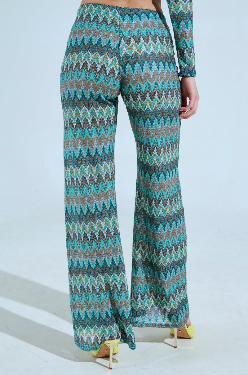 A person models the SHOW ME SOMETHING PANT, featuring a high-rise fit 'n flare chevron pattern in green, blue, and brown. They stand with their back to the camera against a plain background, paired with light yellow high heels. The stretchy knit fabric offers style and comfort. 3