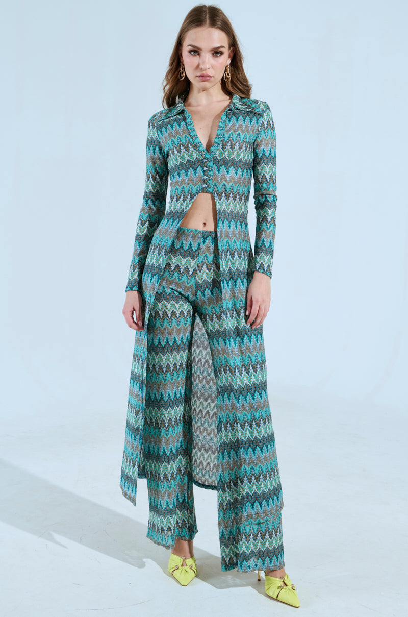 A woman confidently wears the SHOW ME SOMETHING PANT, a high-rise fit 'n flare outfit with teal and blue chevron patterns, stretchy knit fabric, a front cutout, and long flowing pants. She pairs it with yellow pointed shoes against a light blue background. 4