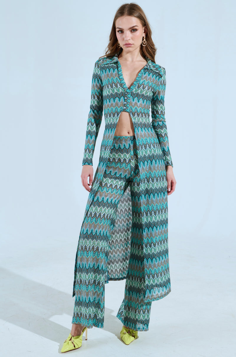 In a studio, a woman models the SHOW ME SOMETHING PANT set, featuring matching wide-leg pants and an open-front long-sleeve V-neck top with a chevron pattern in blue and green shades. She pairs the outfit with yellow pointed shoes on a light blue background. 5