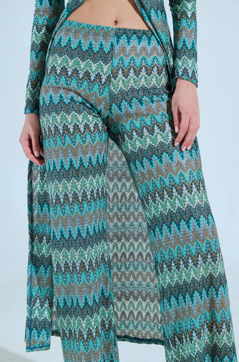 A person shows off the SHOW ME SOMETHING PANT, a vibrant chevron-patterned outfit with stretchy knit fabric. It features high-rise wide-leg pants and a matching long-sleeve top in green, blue, and brown hues on a simple light gray backdrop. 6