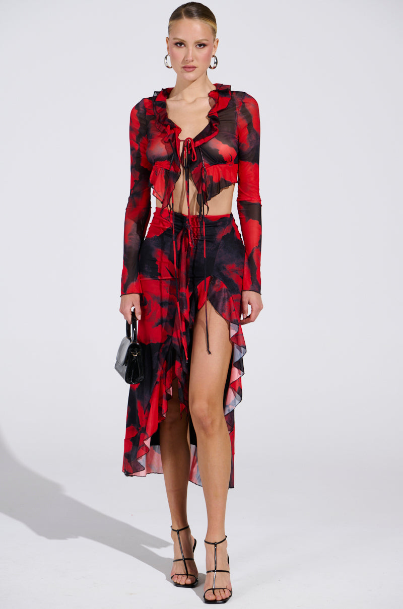 A person in a red and black floral two-piece outfit with ruched detailing, showcasing a long-sleeved top with a CAROLINA MESH MIDI SKIRT featuring a high-slit. They're accessorized with a small black bag, hoop earrings, and strappy heels against a plain background.
