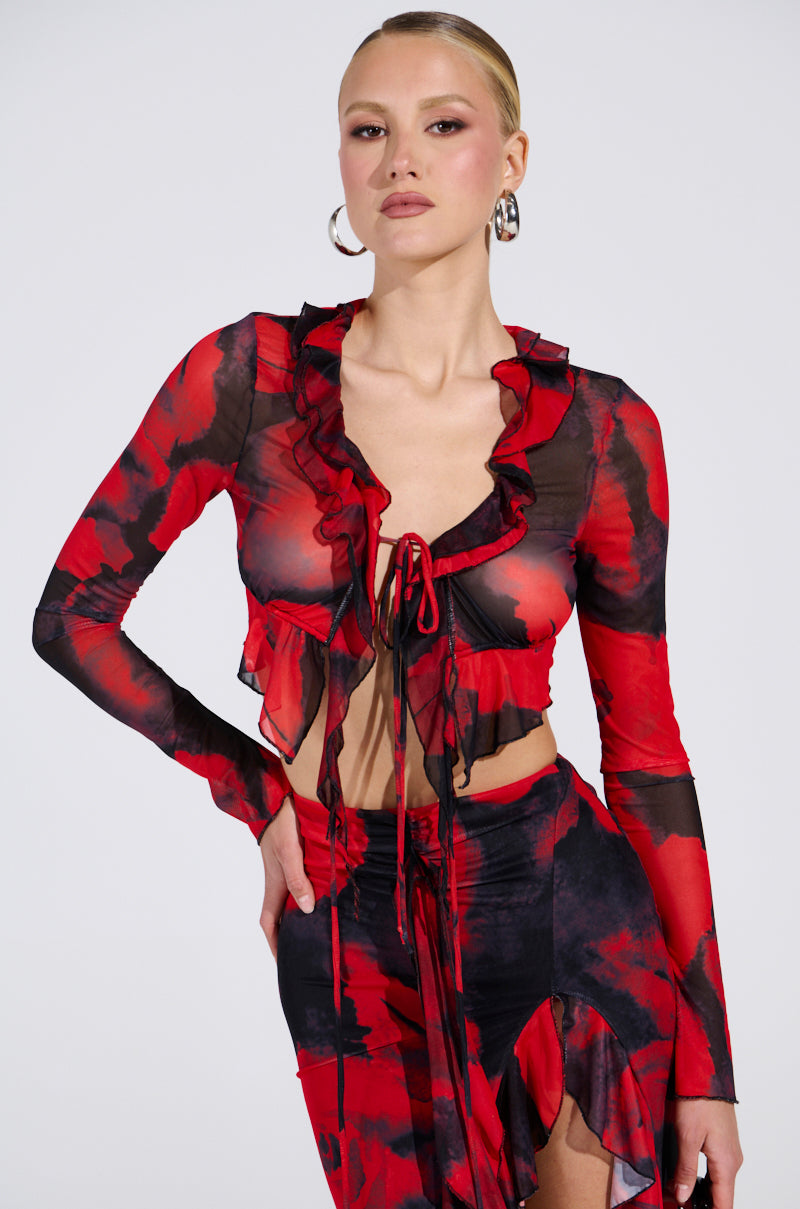 Wearing the CAROLINA LONG SLEEVE MESH TOP, a sheer ruffled red and black piece with an abstract pattern, paired with a matching skirt, they pose confidently. Adorned with hoop earrings and a sleek hairstyle, they stand against a plain white background.