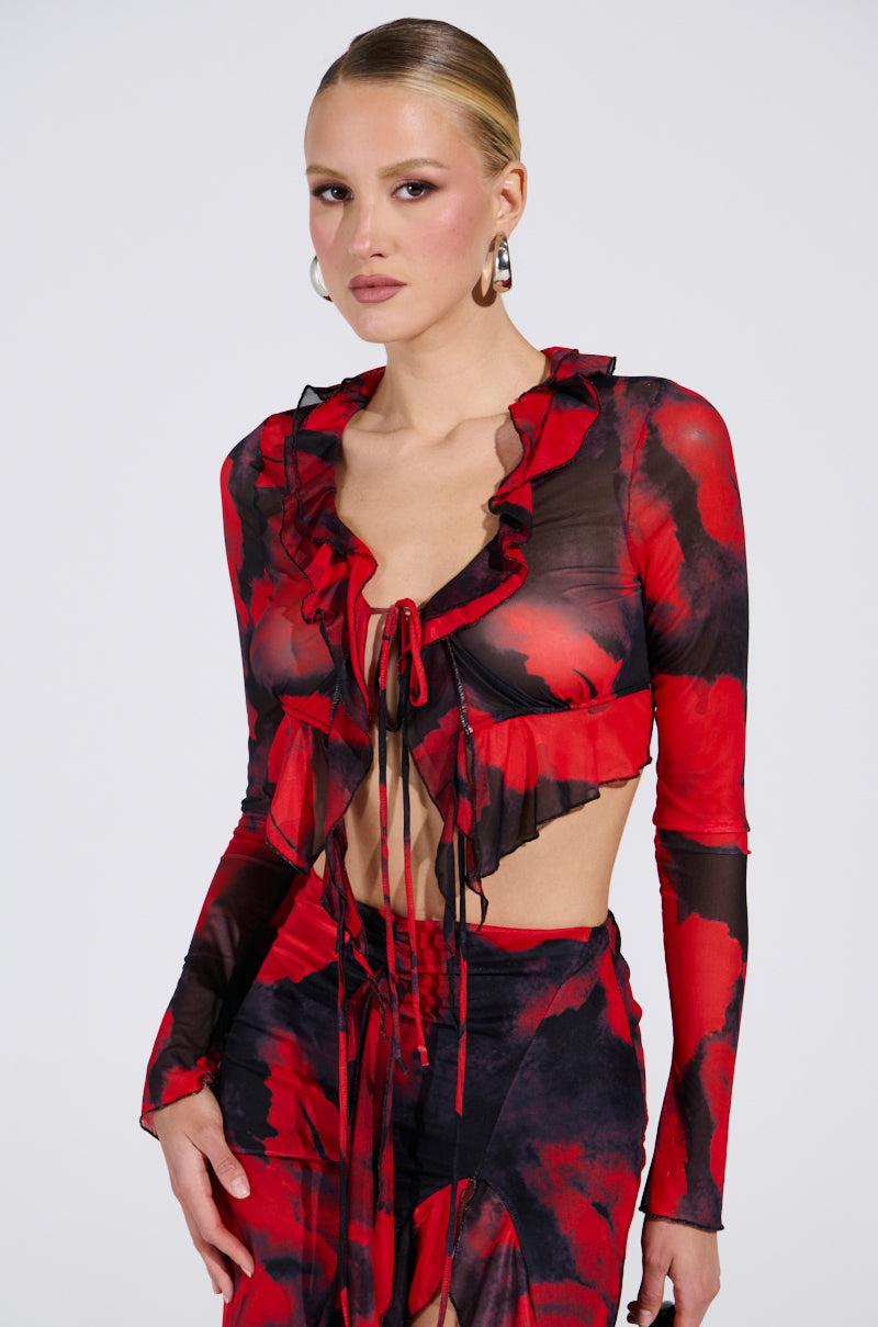 A woman confidently poses against a plain backdrop wearing the CAROLINA LONG SLEEVE MESH TOP, a red and black sheer, ruffled piece with abstract patterns, paired with a matching skirt. She has slicked-back hair and wears hoop earrings.