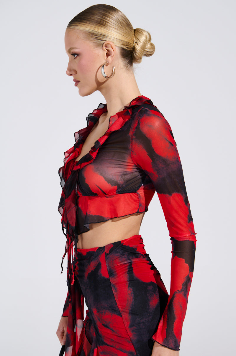 A person with blonde hair in a bun wears the CAROLINA LONG SLEEVE MESH TOP, a red and black ruffled outfit with an abstract pattern. The cropped silhouette shows them standing sideways against a plain white background.