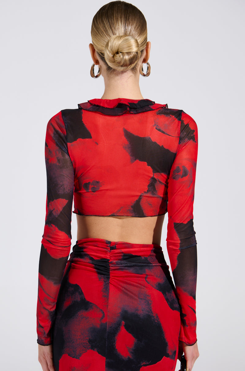 Rear view of a blonde-haired person in a sleek bun, wearing the CAROLINA LONG SLEEVE MESH TOP, boasting a red and black abstract floral pattern. The cropped silhouette pairs beautifully with the high-waisted skirt. They face away from the camera.